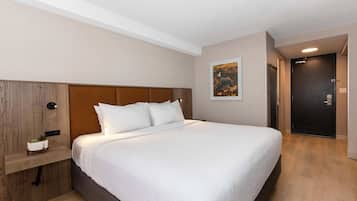 Una habitación de hotel moderna con una cama grande, un cabecero de madera, una mesita de noche con una planta y una puerta que da a otra habitación.