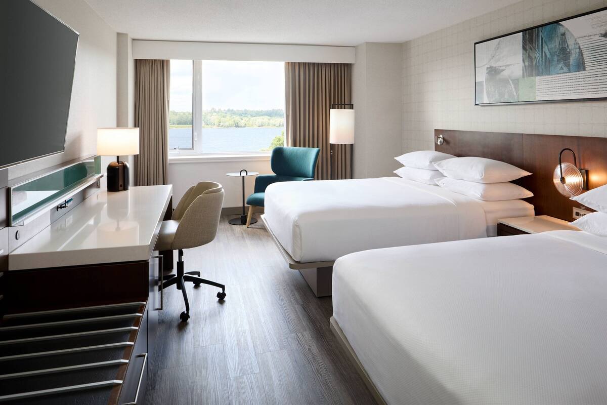 Quarto de hotel com duas camas, uma escrivaninha com computador, uma cadeira, uma televisão e janela com vista para o exterior.
