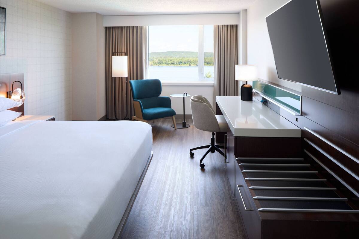 Quarto de hotel com cama, escrivaninha, cadeira, TV e vista para um lago.