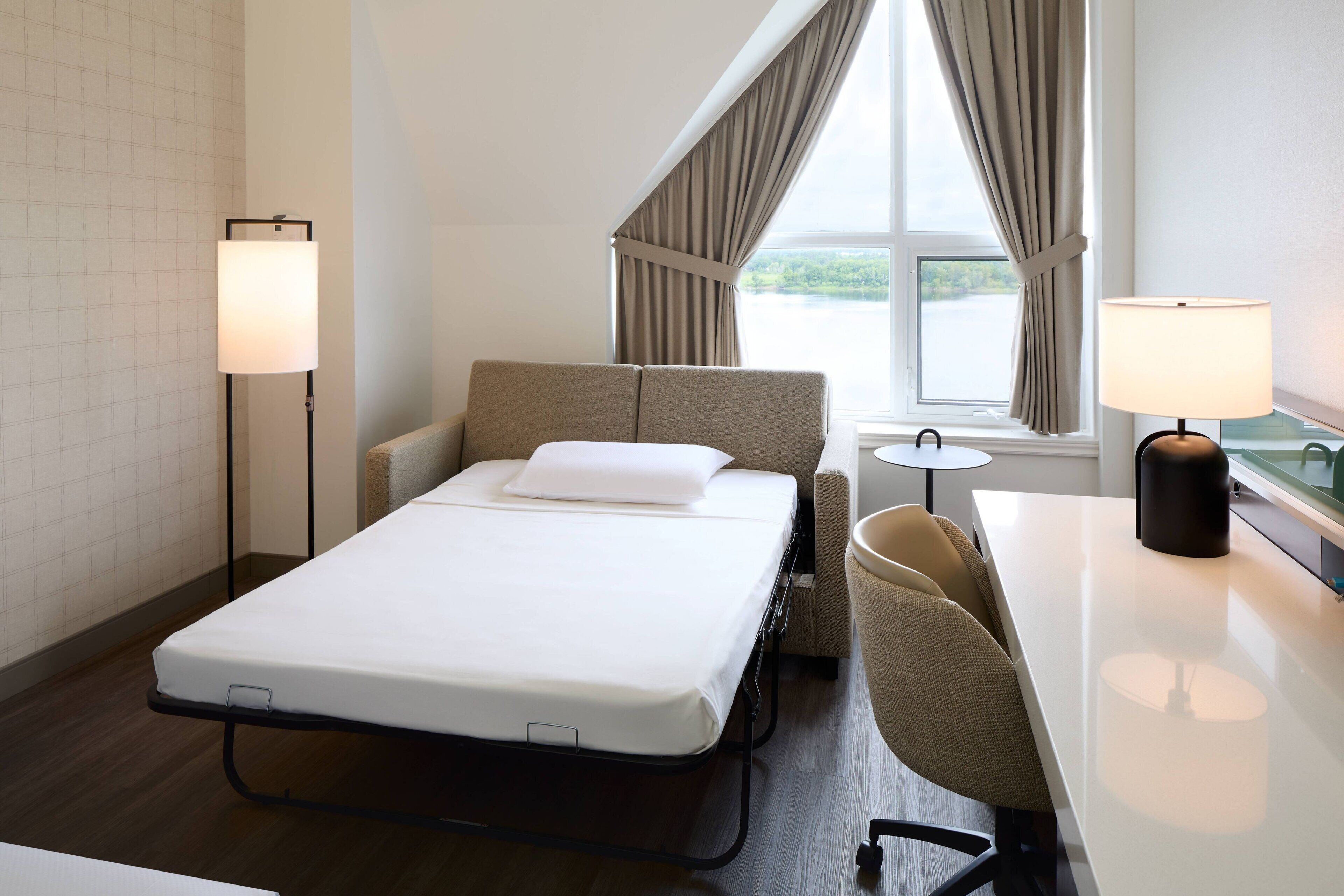 Une chambre d’hôtel avec un lit, un bureau, deux lampes et une fenêtre avec des rideaux.