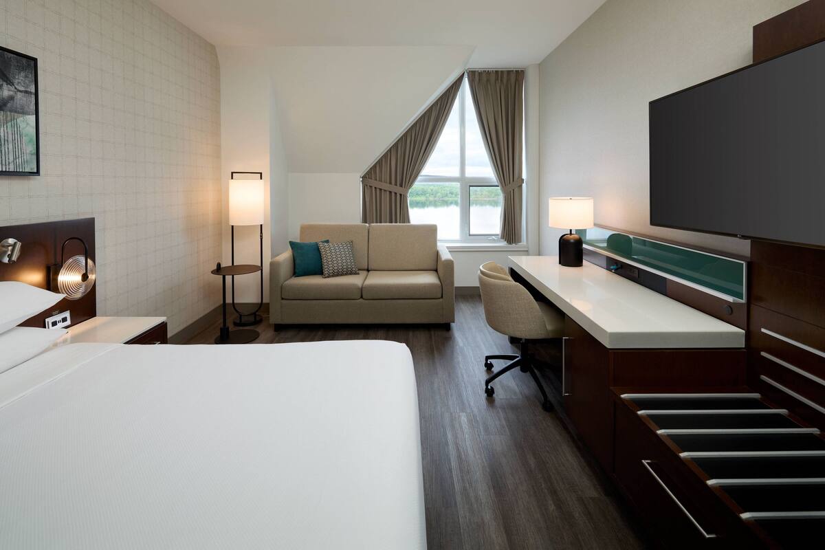 Quarto de hotel com uma cama grande, um sofá, uma escrivaninha com televisão de tela plana, uma luminária e vista para o exterior.