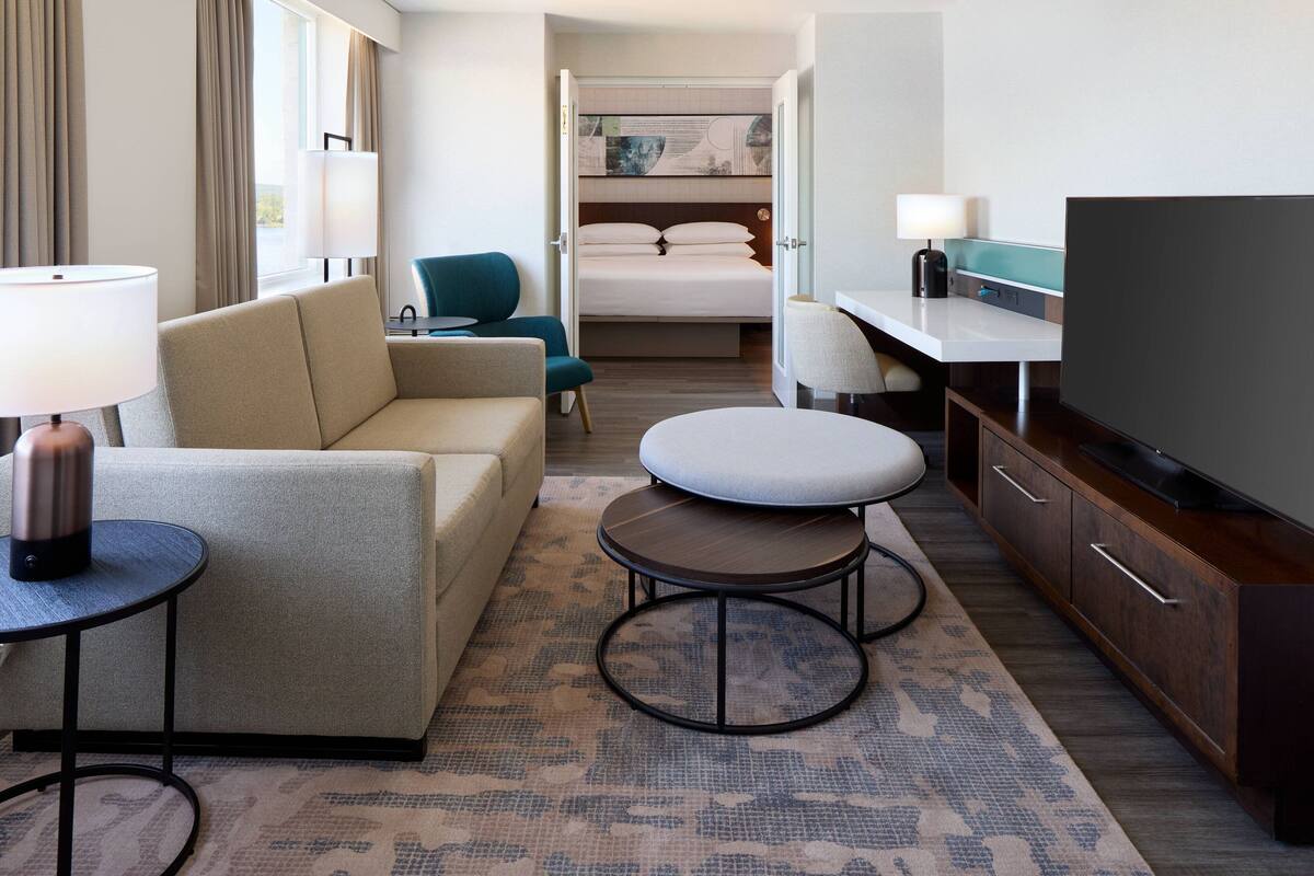 Quarto de hotel com sofá, mesa redonda pequena, televisão, escrivaninha e cama.
