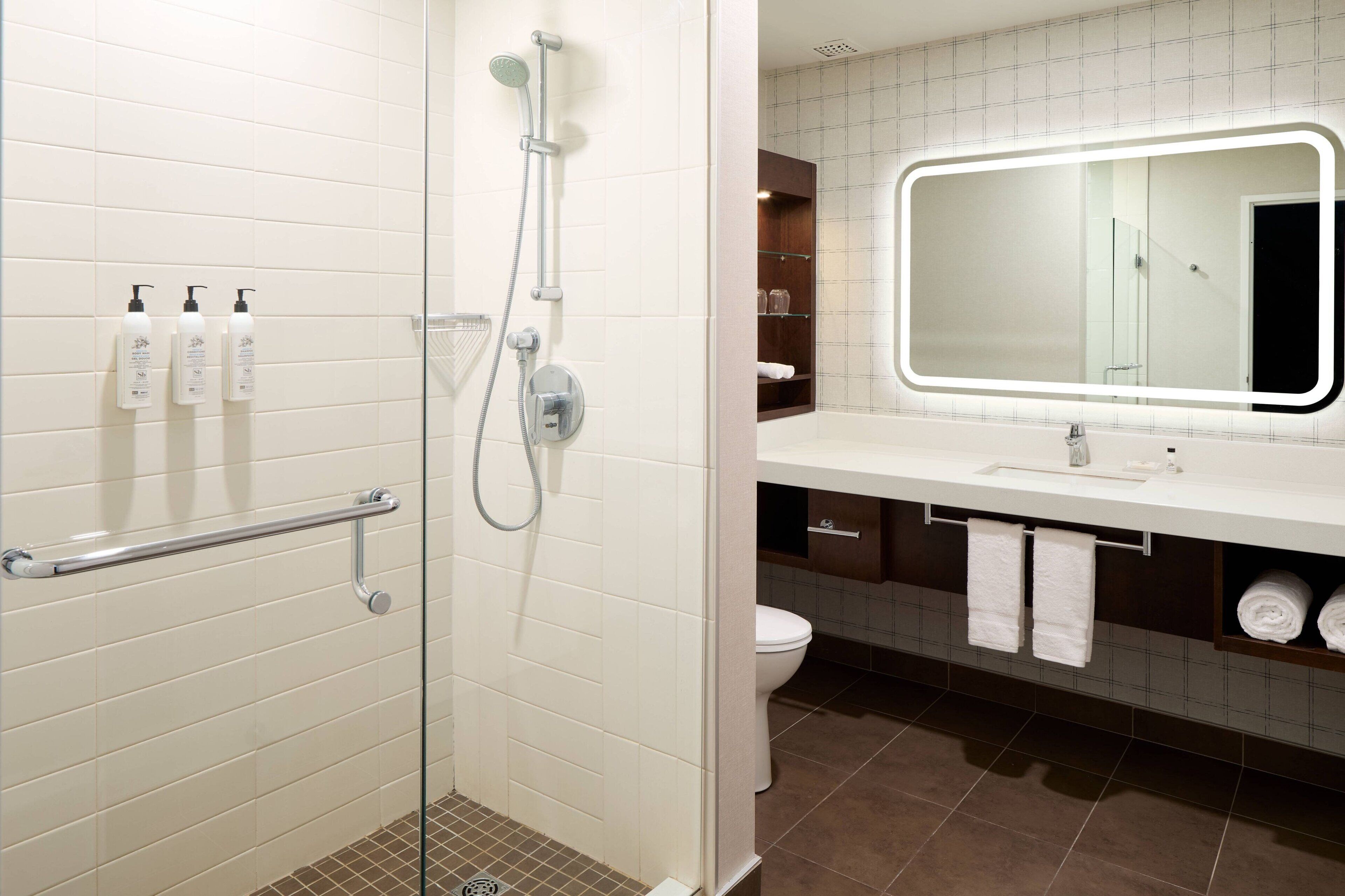 A modern bathroom with a glass-enclosed shower, white tiled walls, a large mirror with LED lighting, and a white sink with a towel rack.