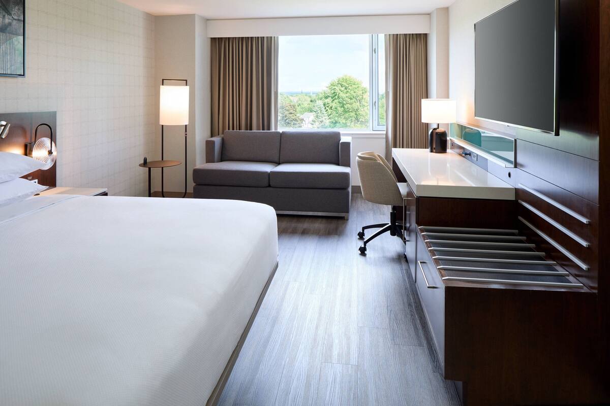 Quarto de hotel com uma cama grande, um sofá, uma escrivaninha com cadeira, uma televisão de tela plana e vista para o exterior.
