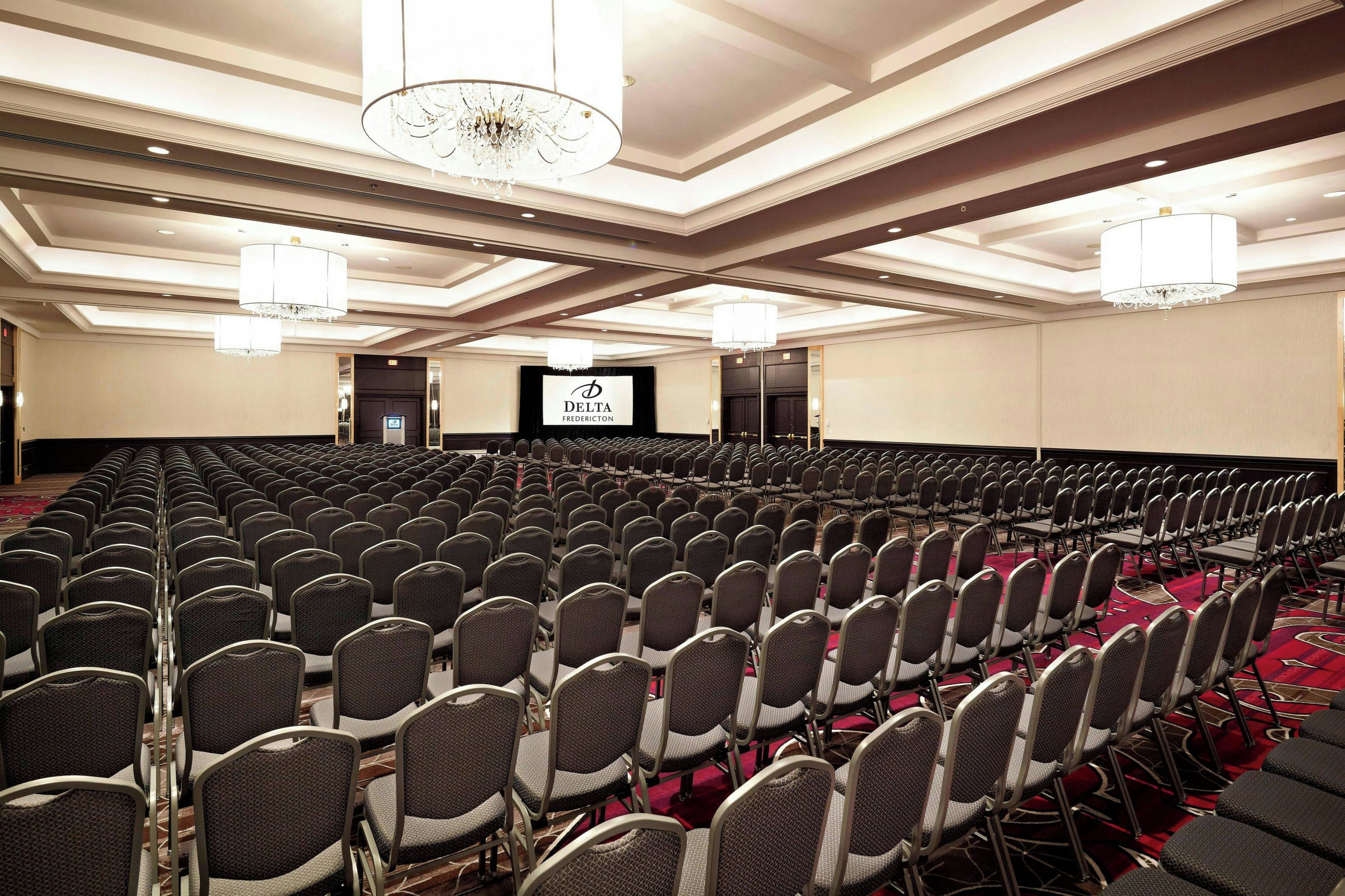 Une grande salle de conférence avec des rangées de chaises, une scène dotée d’un écran et des lustres.