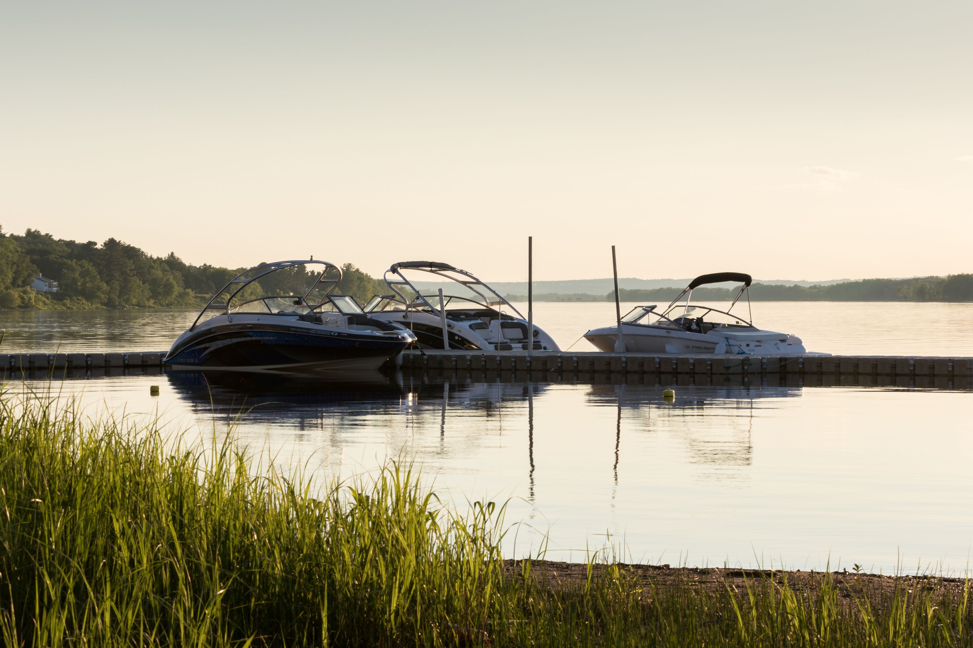 Two boats docked at a marina with calm water and a forested shoreline in the background.