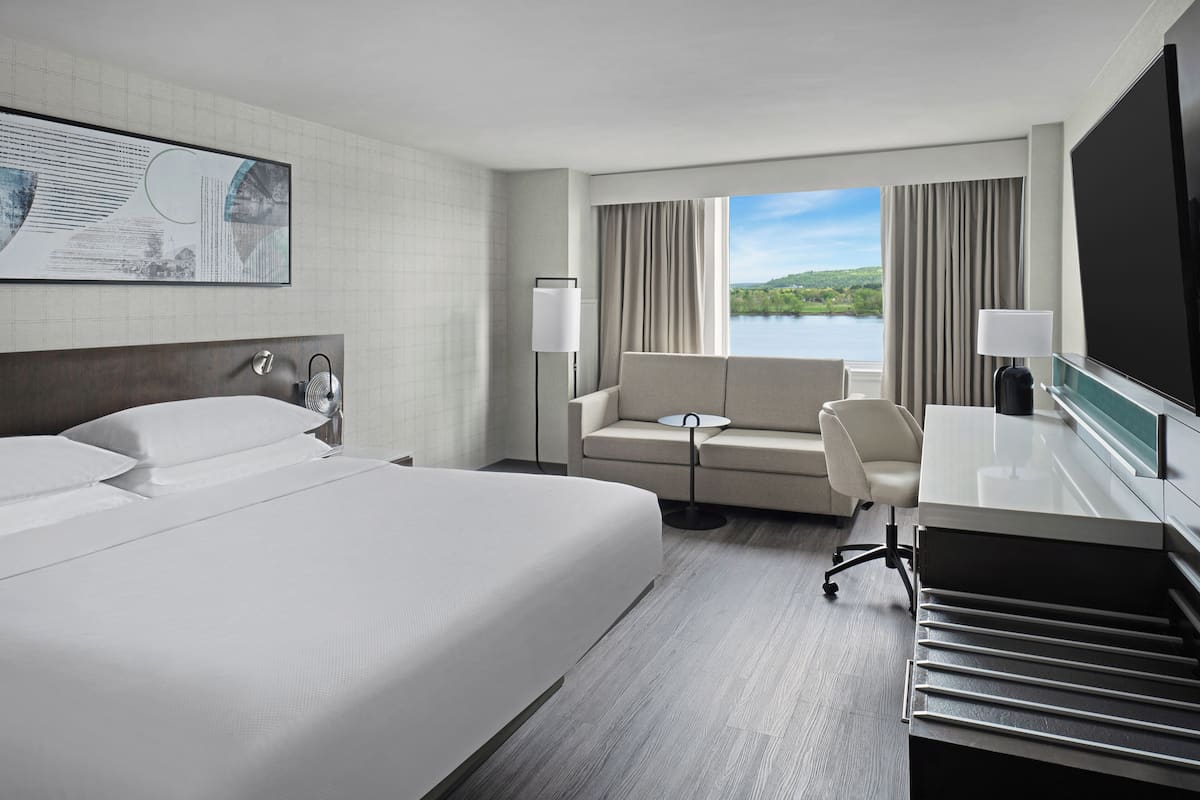 Quarto de hotel com uma cama grande, um sofá, uma escrivaninha e vista para um lago.