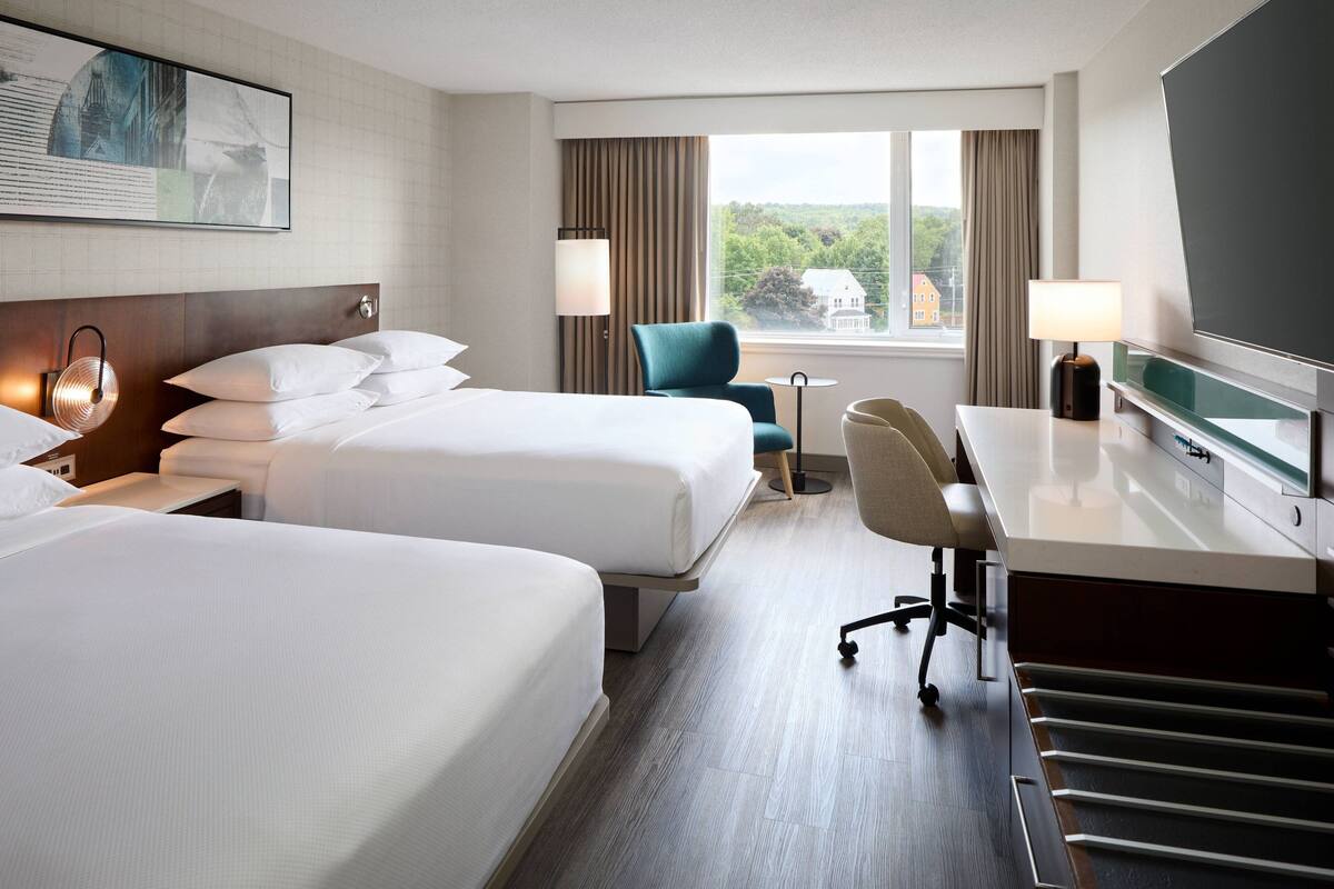 Quarto de hotel com duas camas, uma escrivaninha, uma cadeira, televisão e janela com vista para áreas verdes e edifícios.