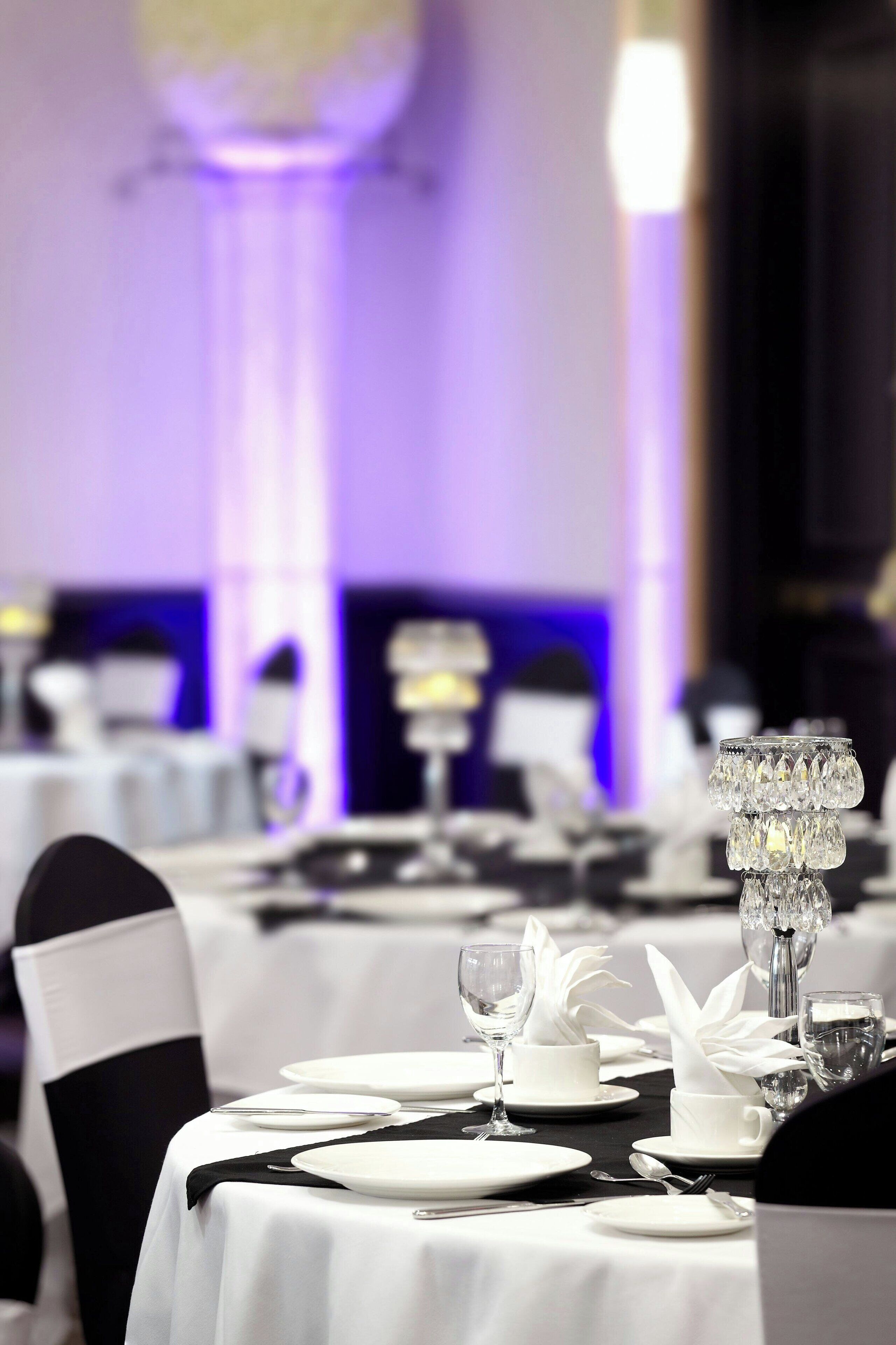 Un salón de banquetes con mesas dispuestas para un evento formal, con manteles blancos, centros de mesa de cristal y cintas negras en las sillas.