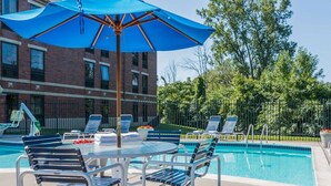 A poolside area with a table and chairs, a blue umbrella, and lounge chairs around a pool. - Quality Inn Auburn Hills - Detroit North (Auburn Hills)