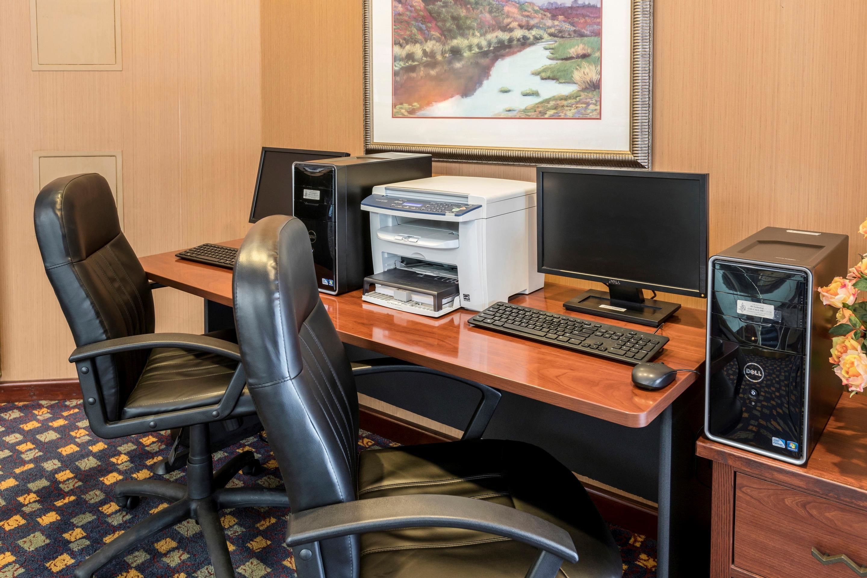 A hotel office with two black leather chairs, a wooden desk, a desktop computer, a printer, and a framed landscape painting on the wall.