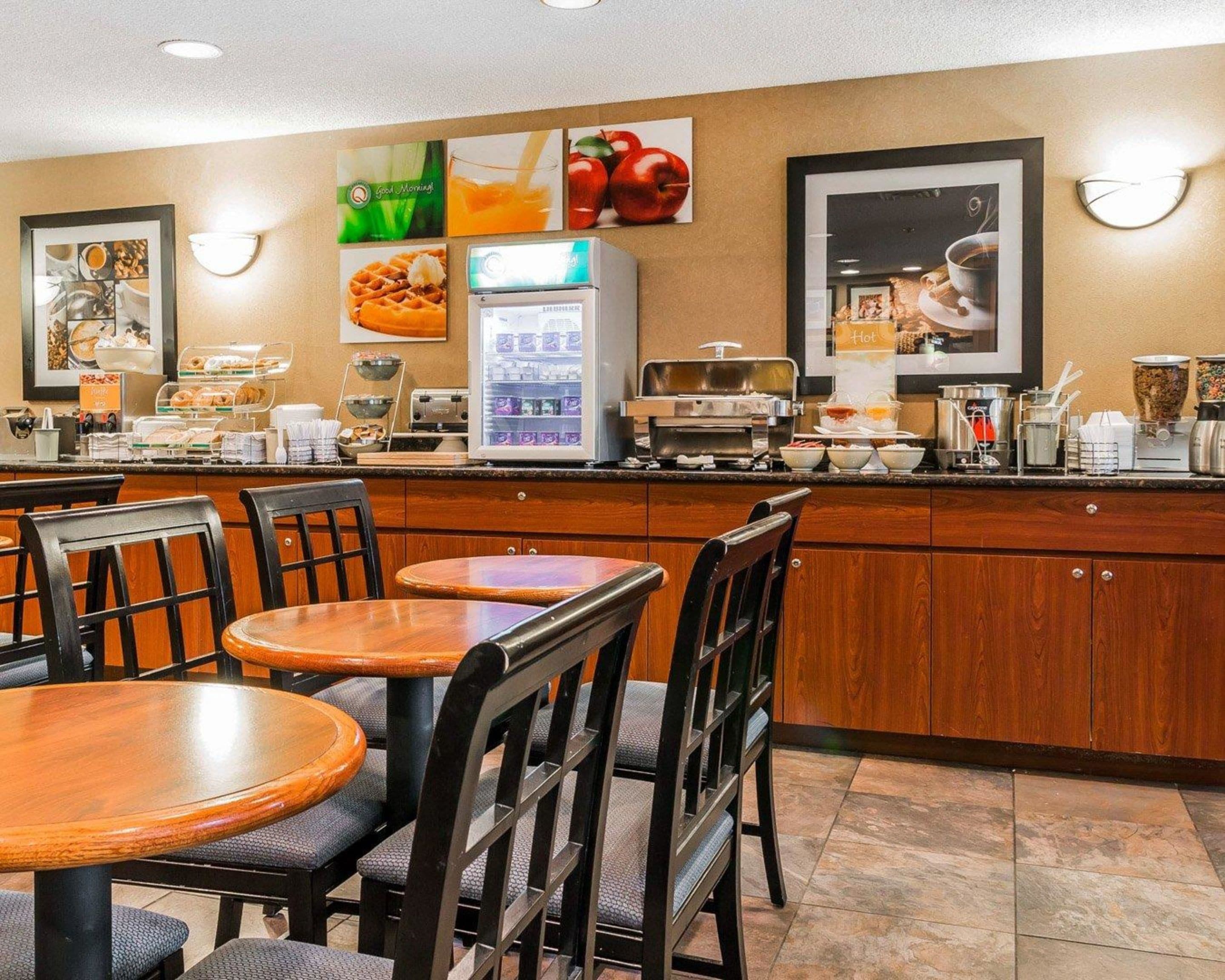 A breakfast area with wooden tables and chairs, a refrigerator with beverages, and a buffet counter with various food items.