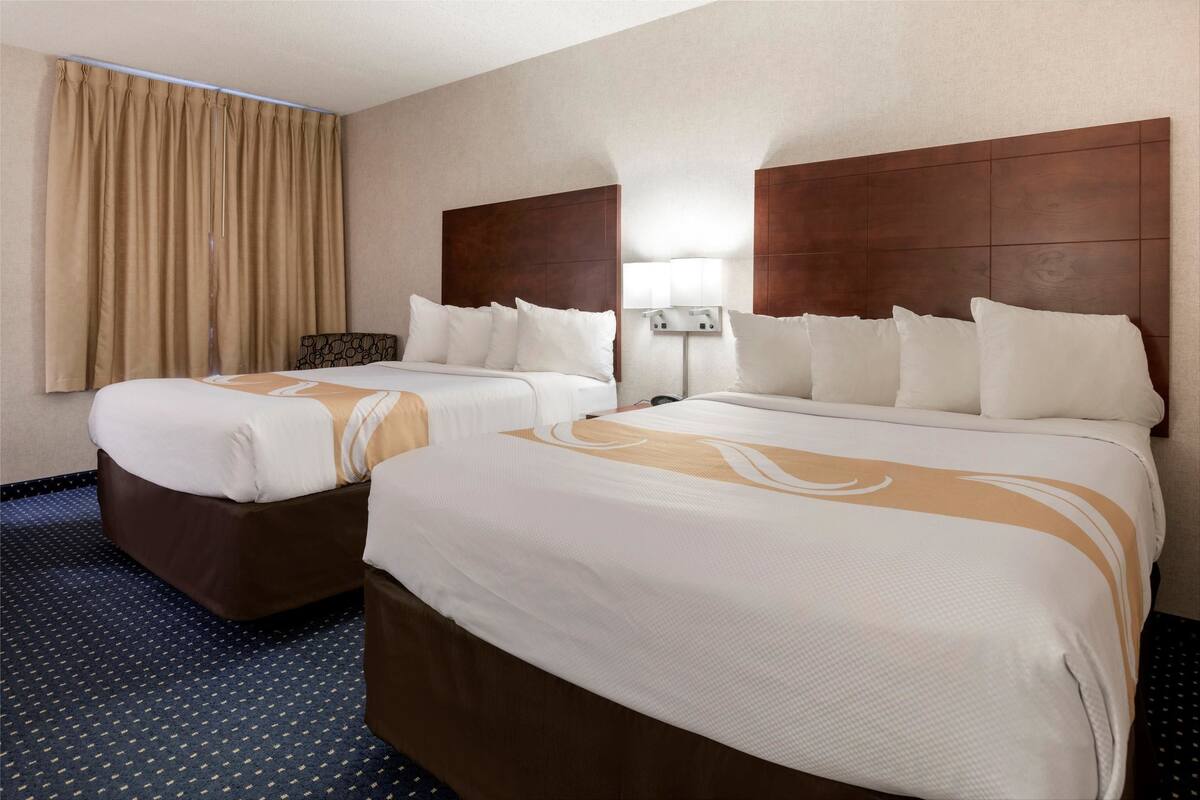 A hotel room with two beds, a large window with curtains, a wall-mounted lamp, and a carpeted floor.
