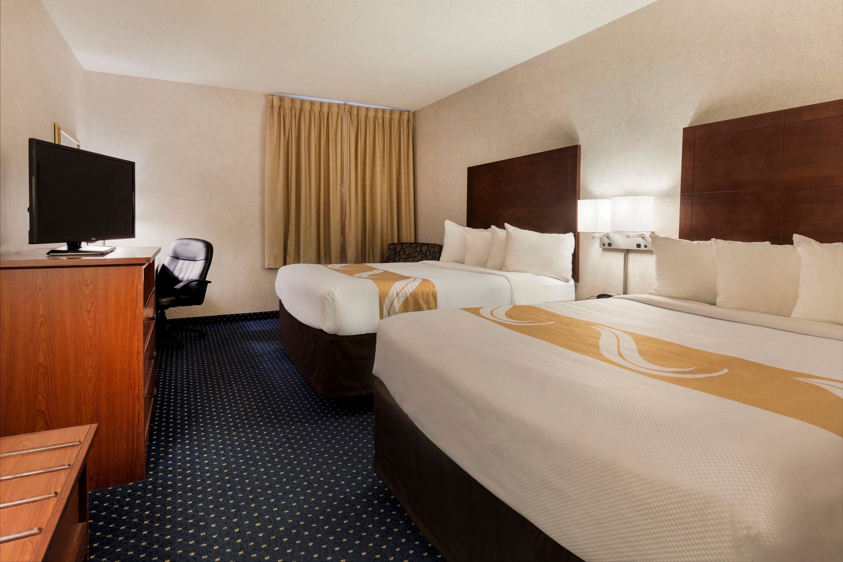 Ein Hotelzimmer mit zwei Betten, einem Schreibtisch mit Computer, einem Sessel und einem großen Fenster mit Vorhängen.