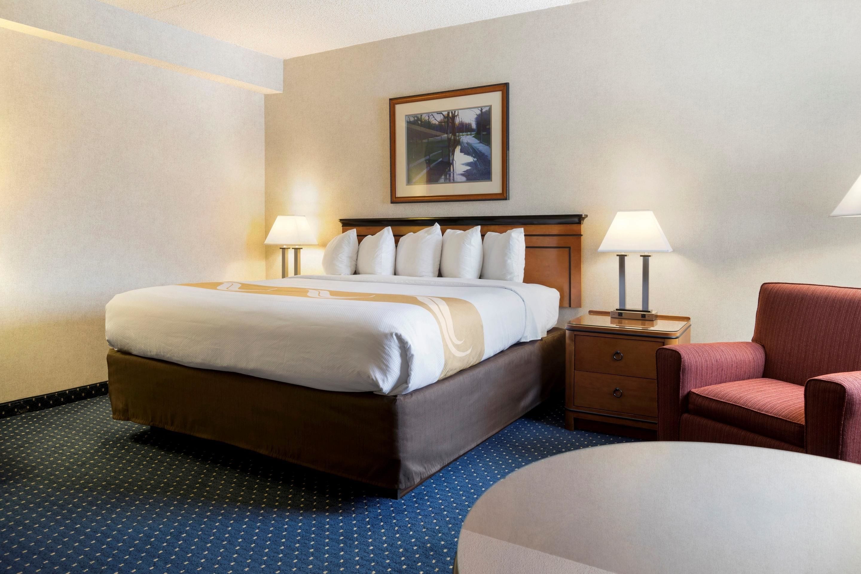 A hotel room with a large bed, two bedside tables with lamps, a chair, and a framed picture on the wall.