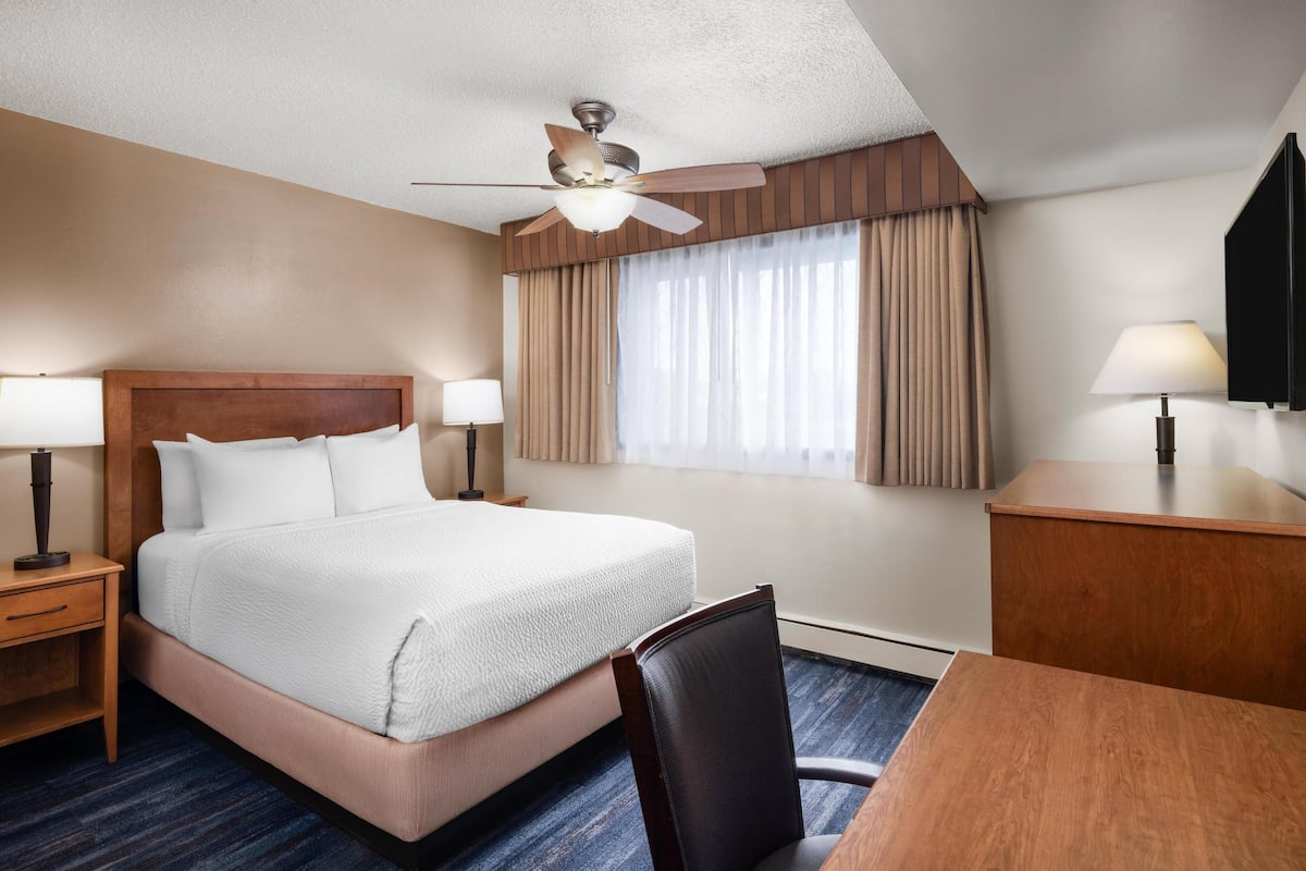 Habitación de hotel con una cama grande, dos mesitas de noche con lámparas, un escritorio con silla, un ventilador de techo y una ventana con cortinas.