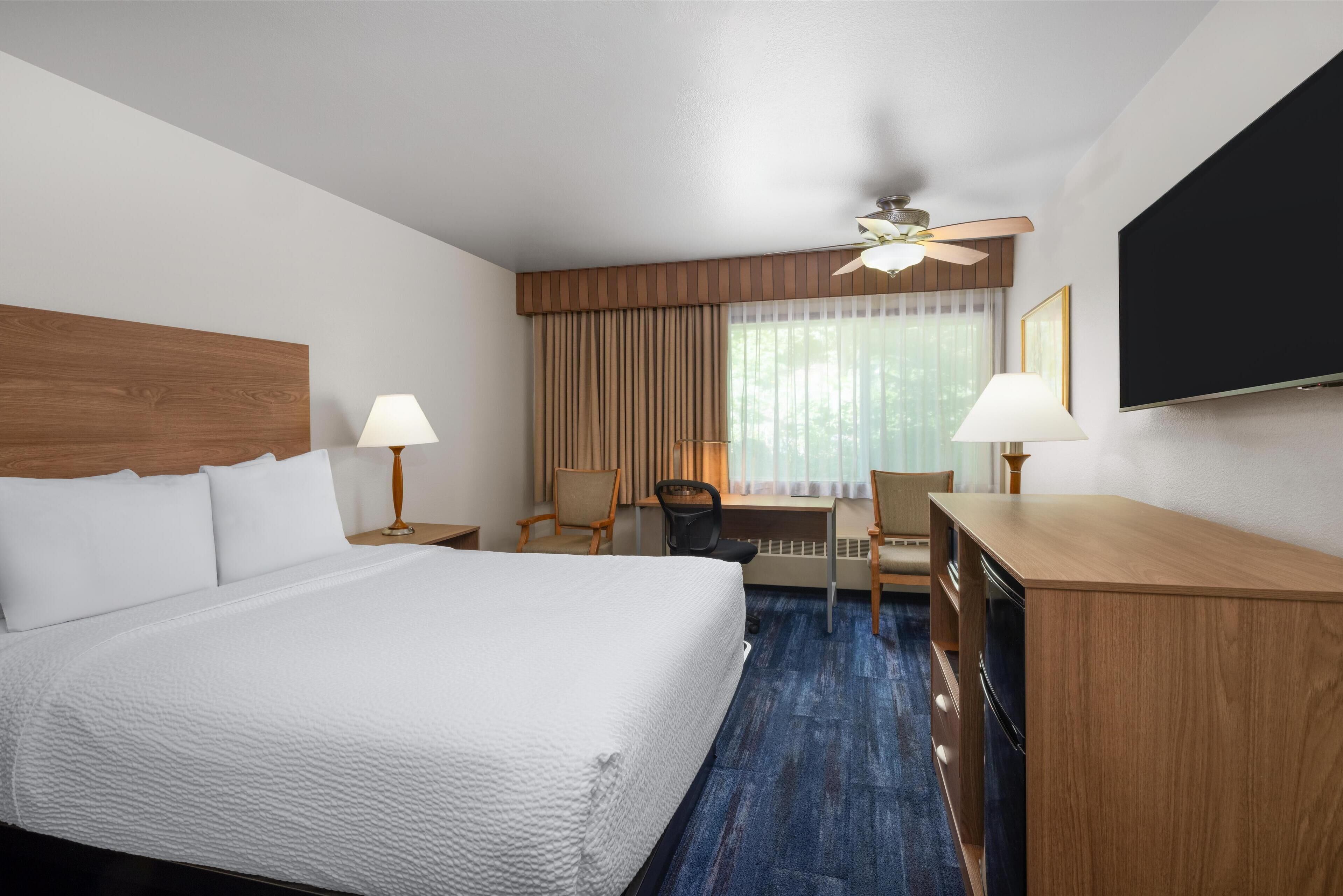 A hotel room with a large bed, a desk, two chairs, a ceiling fan, and a flat-screen TV.