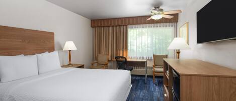 A hotel room with a large bed, a desk, two chairs, a ceiling fan, and a flat-screen TV.