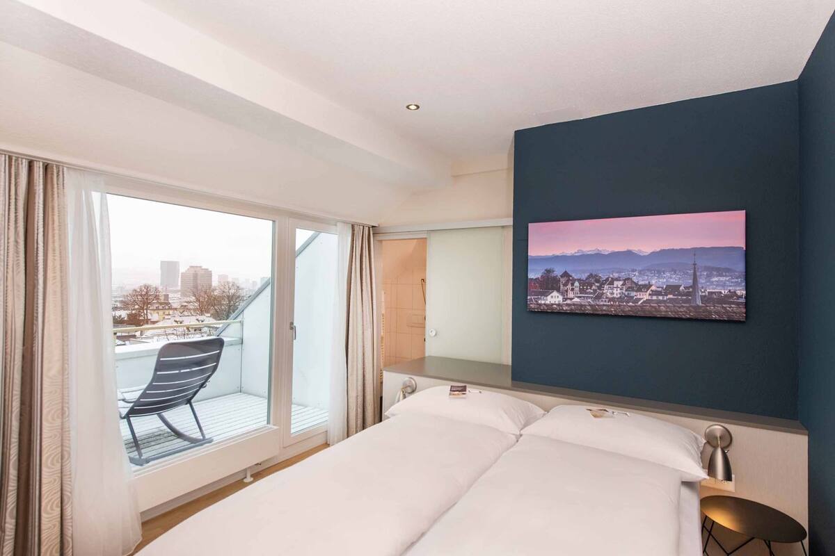 A hotel room with a bed, a chair, a desk, a painting, and a view of a cityscape.