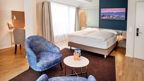 Una habitación de hotel con una cama, un sillón azul, una pequeña mesa redonda con una botella y una caja, y una pintura de un paisaje urbano en la pared.