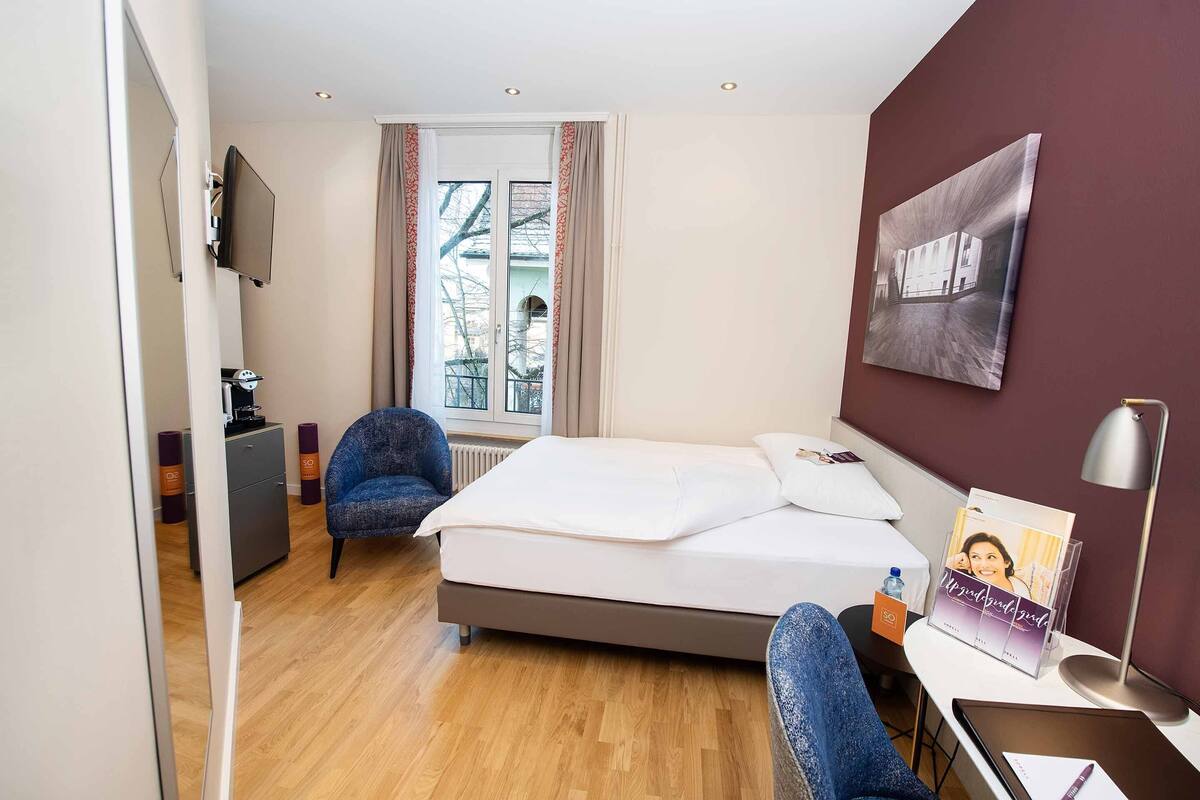 A hotel room with a bed, a desk with a lamp, a chair, a radiator, a window with curtains, and a painting on the wall.