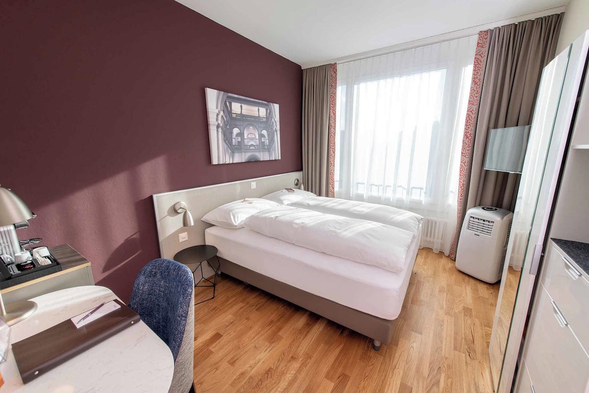 A hotel room with a bed, bedside table, lamp, chair, and a window with curtains.