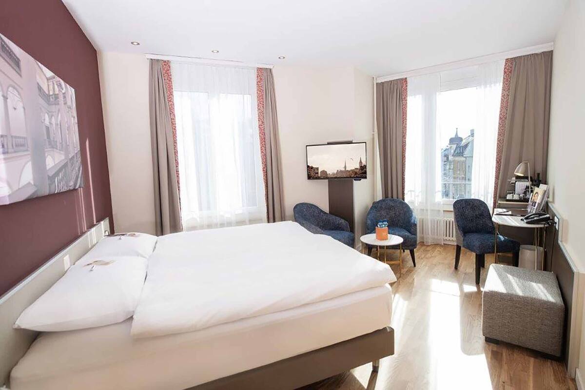 A hotel room with a bed, two armchairs, a small table, and a desk with a lamp.