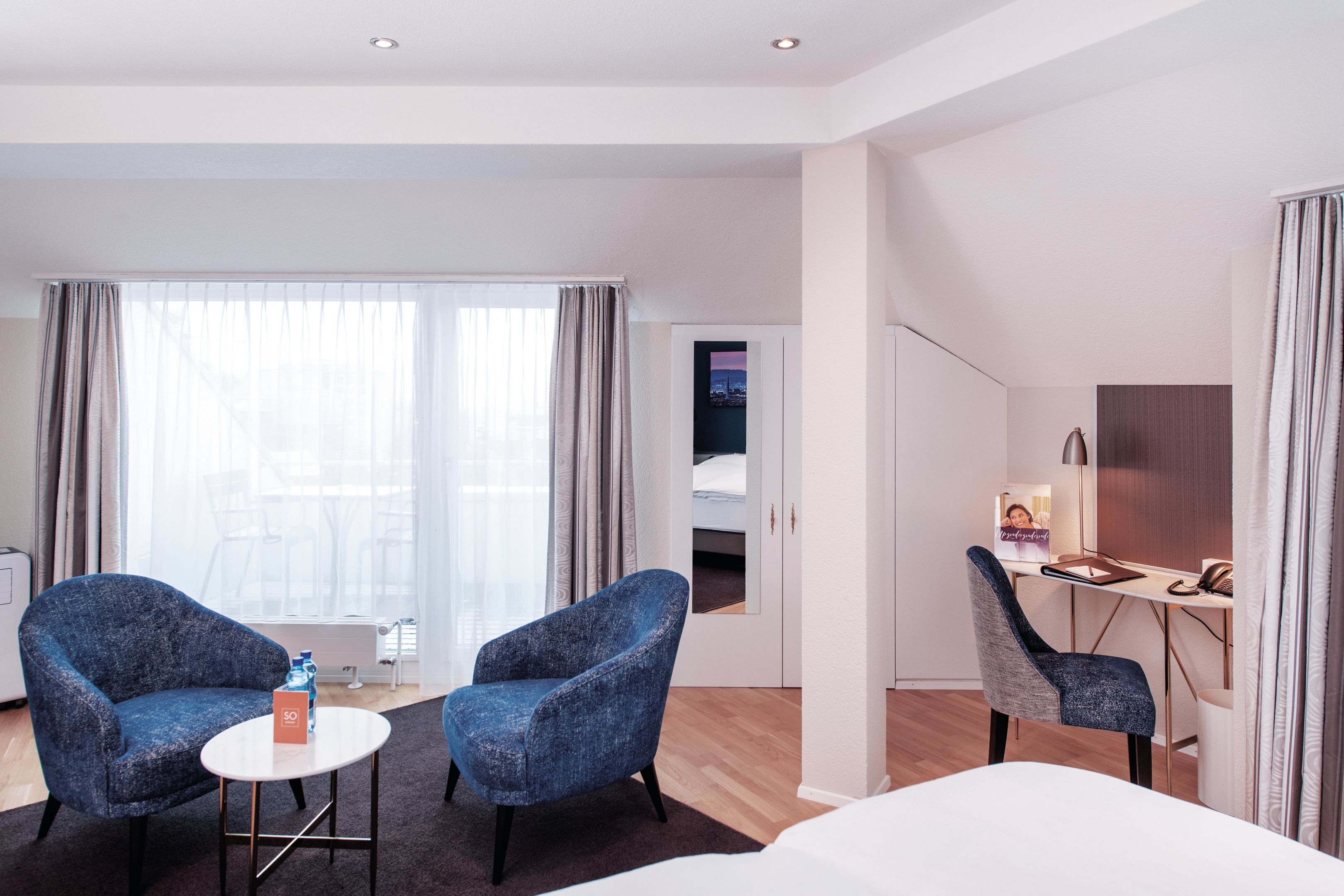 A hotel room with a bed, two armchairs, a desk, and a view of the city through large windows.