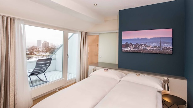 Habitación de hotel con cama, escritorio, silla y un amplio ventanal con vistas a la ciudad.