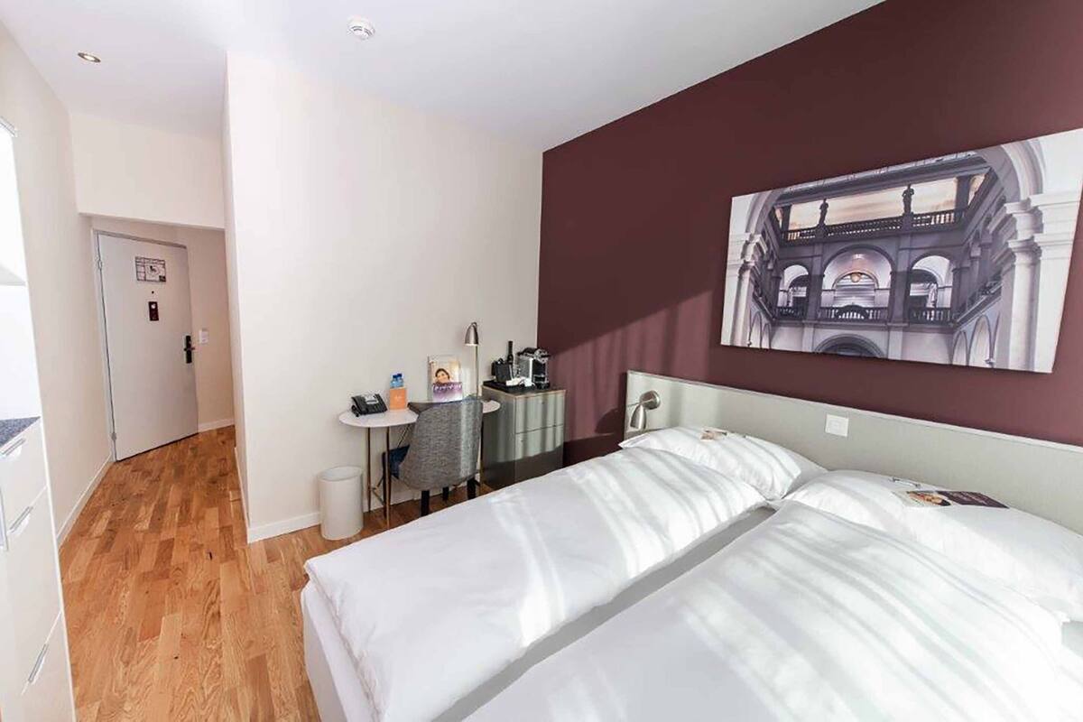 A hotel room with a bed, a desk, a chair, a trash bin, and a wall-mounted picture.