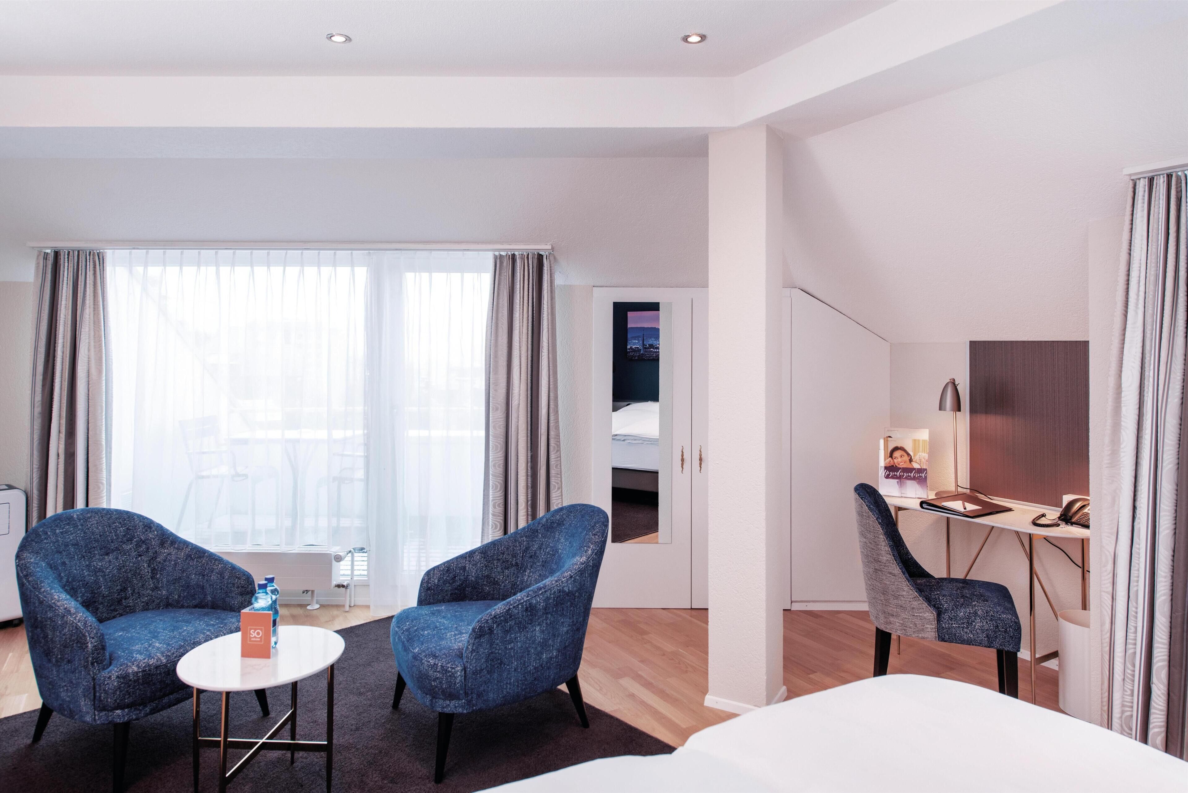 Habitació d'hotel moderna amb un llit, dues butaques, un escriptori amb una làmpada i una finestra gran amb cortines.