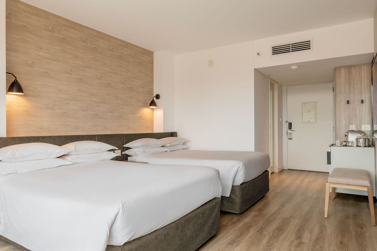 A modern hotel room with two beds, a wooden headboard, a small kitchenette, and a wooden bench.