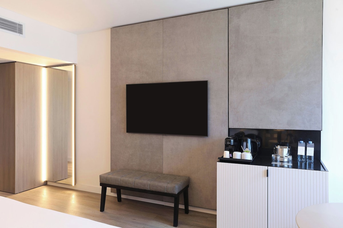 A modern room with a flat-screen TV mounted on a wall, a bench, and a coffee station with a kettle and cups.