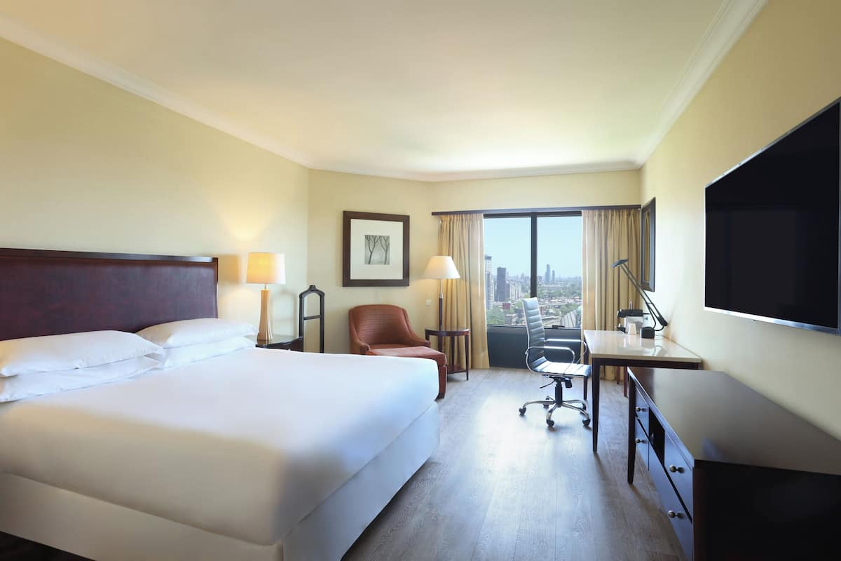 Ein Hotelzimmer mit einem großen Bett, einem Schreibtisch, einem Stuhl, einem Fernseher und einem Fenster mit Blick auf die Stadt.