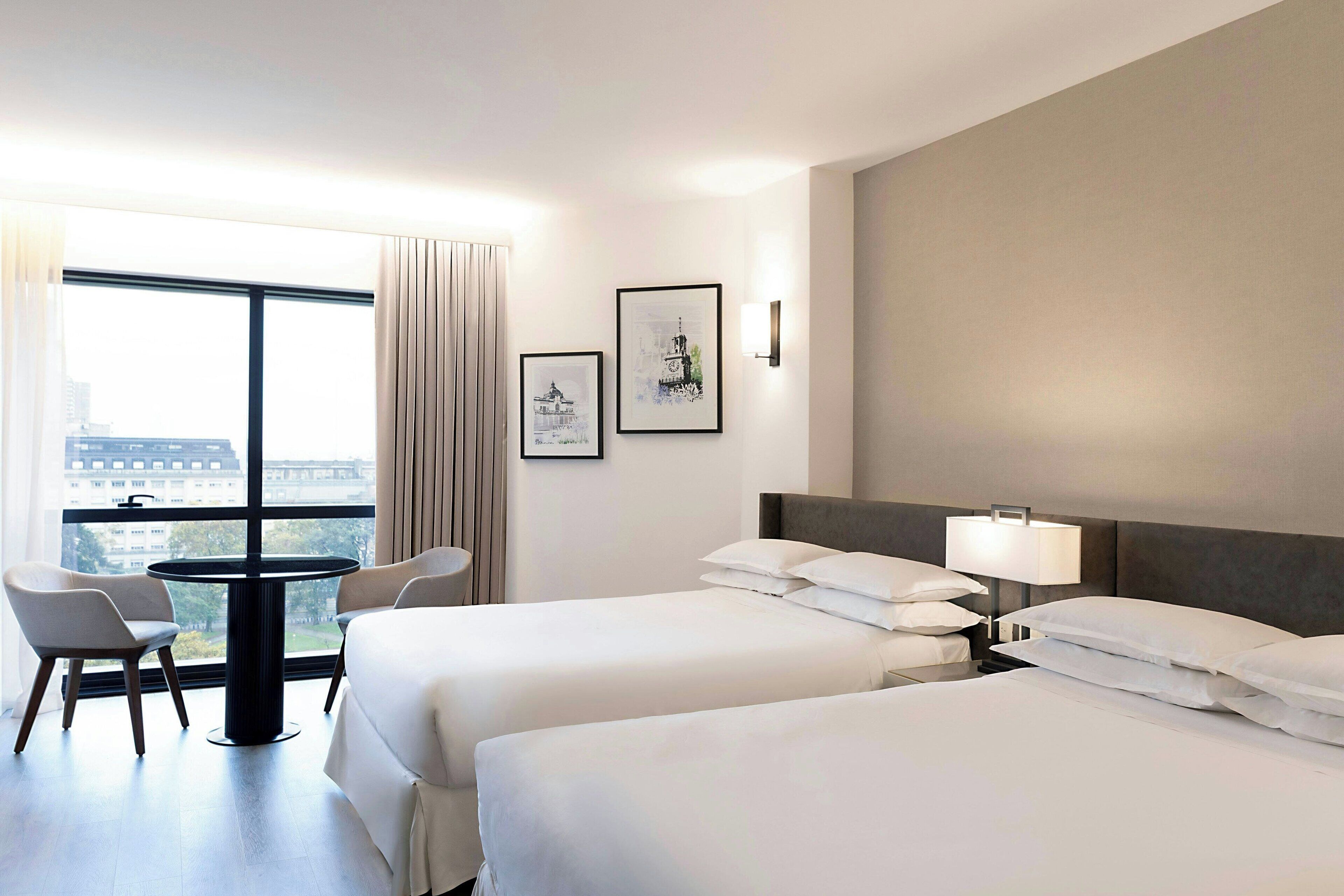 Ein Hotelzimmer mit zwei Betten, einem runden Tisch mit Stühlen, einem großen Fenster mit Blick auf Gebäude und zwei gerahmten Bildern an der Wand.