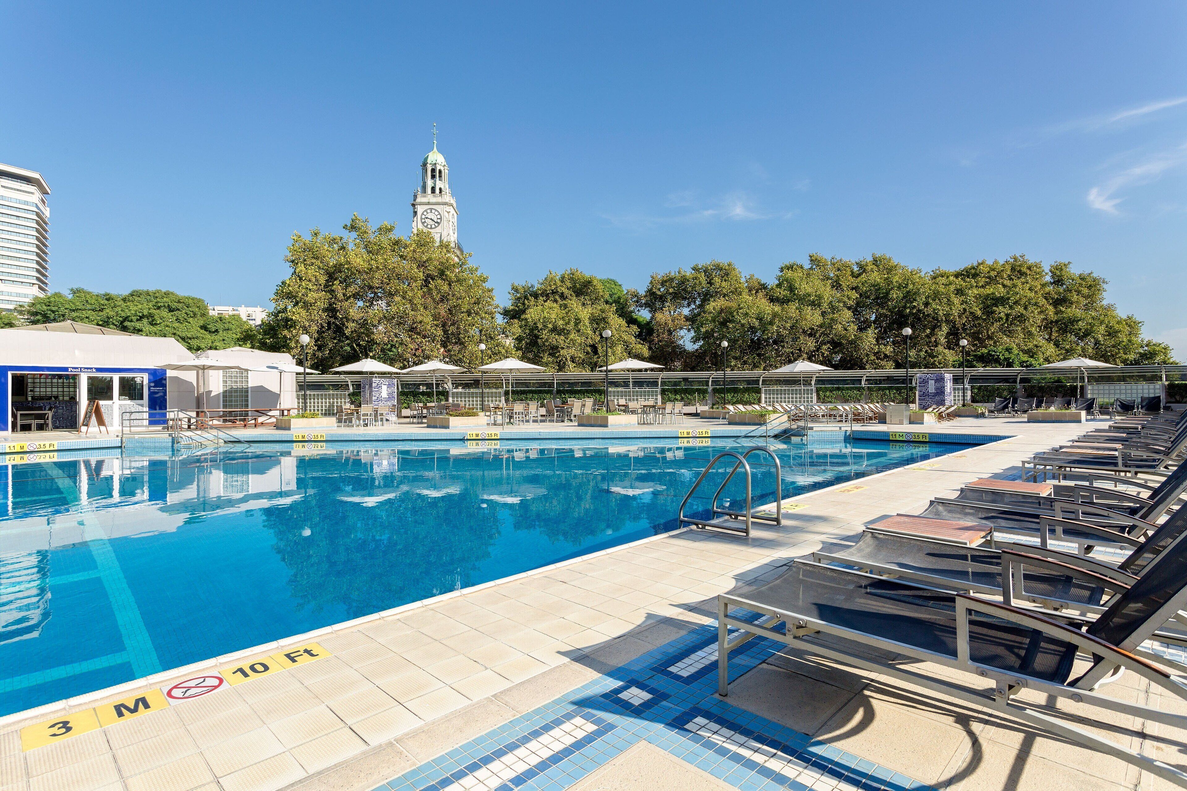 Una piscina con sillones de descanso y sombrillas, una torre del reloj al fondo y un cielo despejado.
