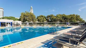 Una piscina con sillones de descanso y sombrillas, una torre del reloj al fondo y un cielo despejado.