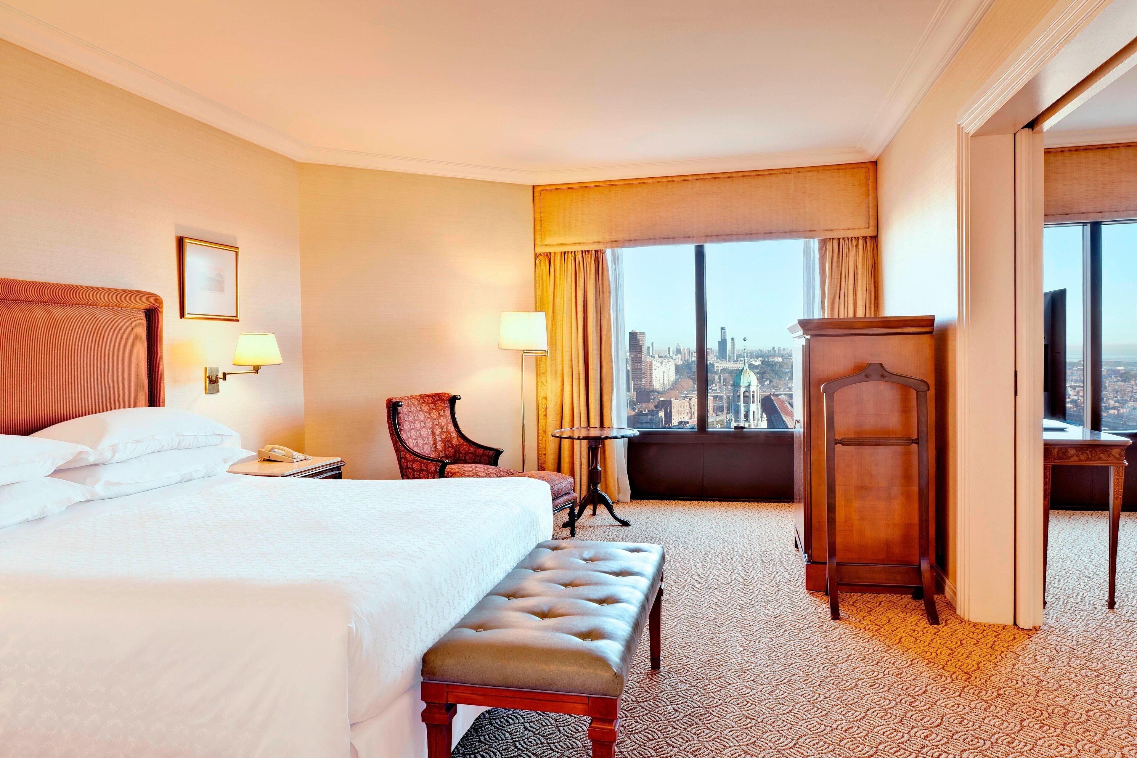 A hotel room with a large bed, a bench, a desk, a chair, and a view of a cityscape through a large window.