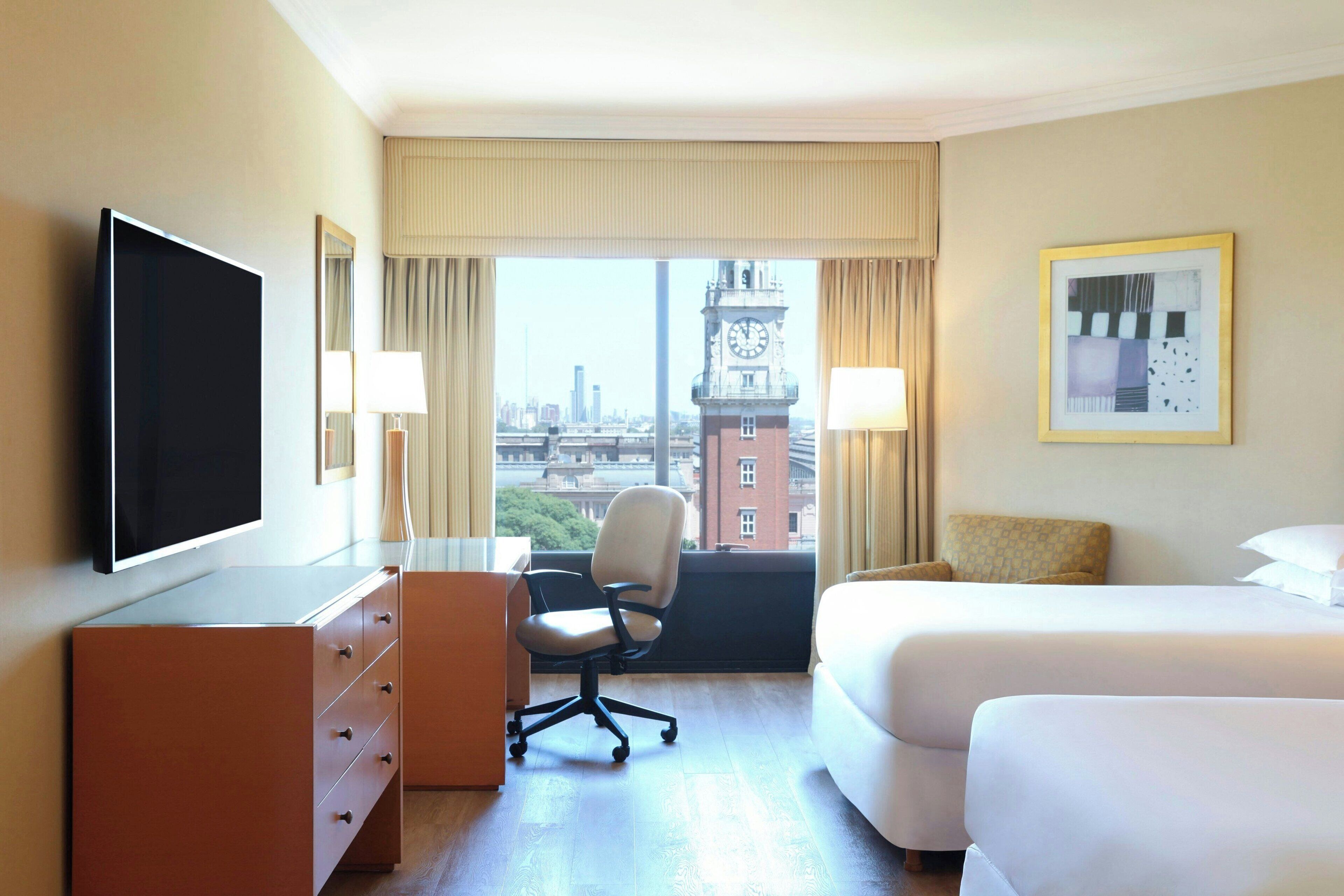 A hotel room with a bed, a desk, a chair, a TV, and a large window with a city view.