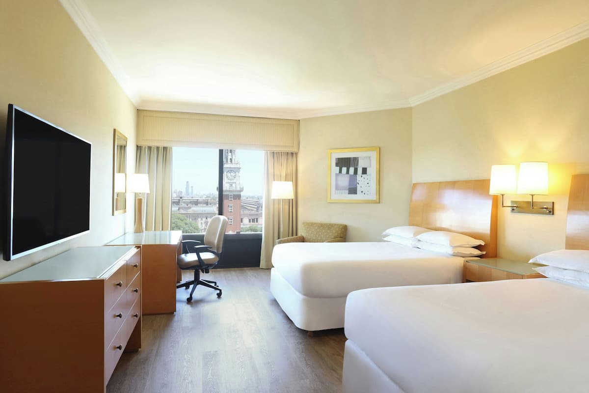 Ein Hotelzimmer mit zwei Betten, einem Schreibtisch, einem Stuhl, einem Fernseher und einem Fenster mit Blick auf die Stadt.