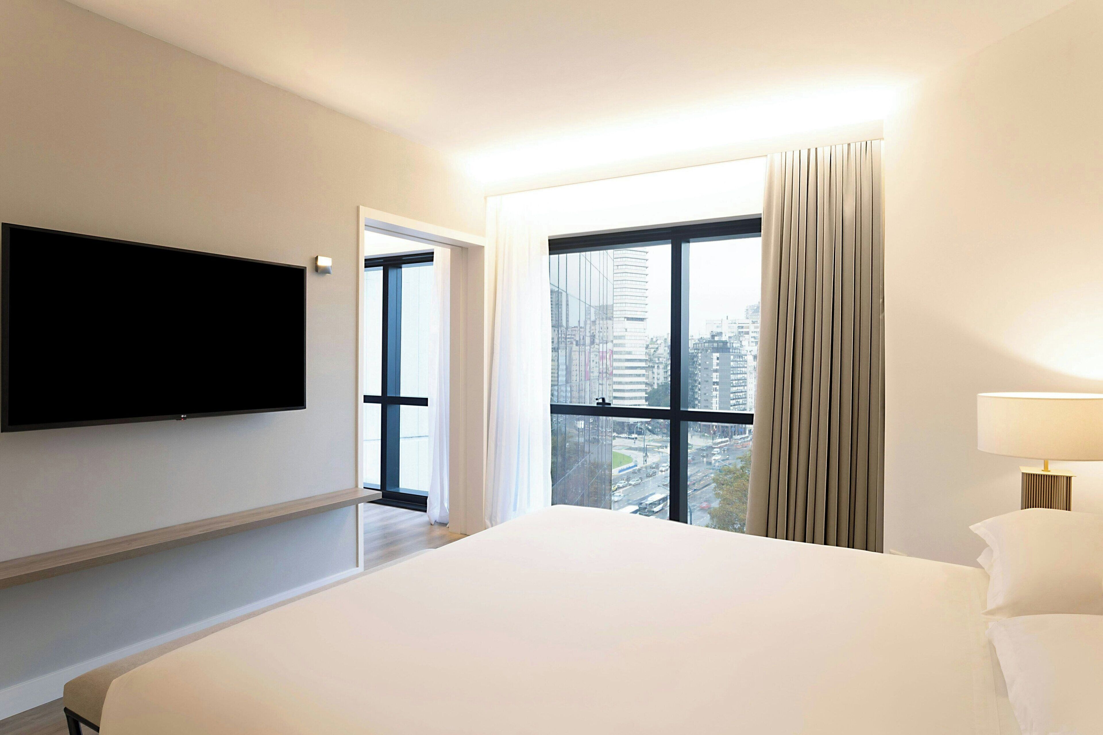 Habitación de hotel con una cama grande, televisión de pantalla plana en la pared, ventana con cortinas y vista a la ciudad.