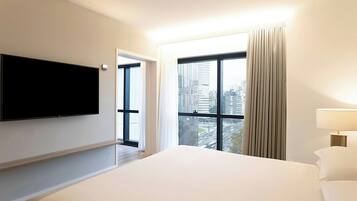 Habitación de hotel con una cama grande, televisión de pantalla plana en la pared, ventana con cortinas y vista a la ciudad.