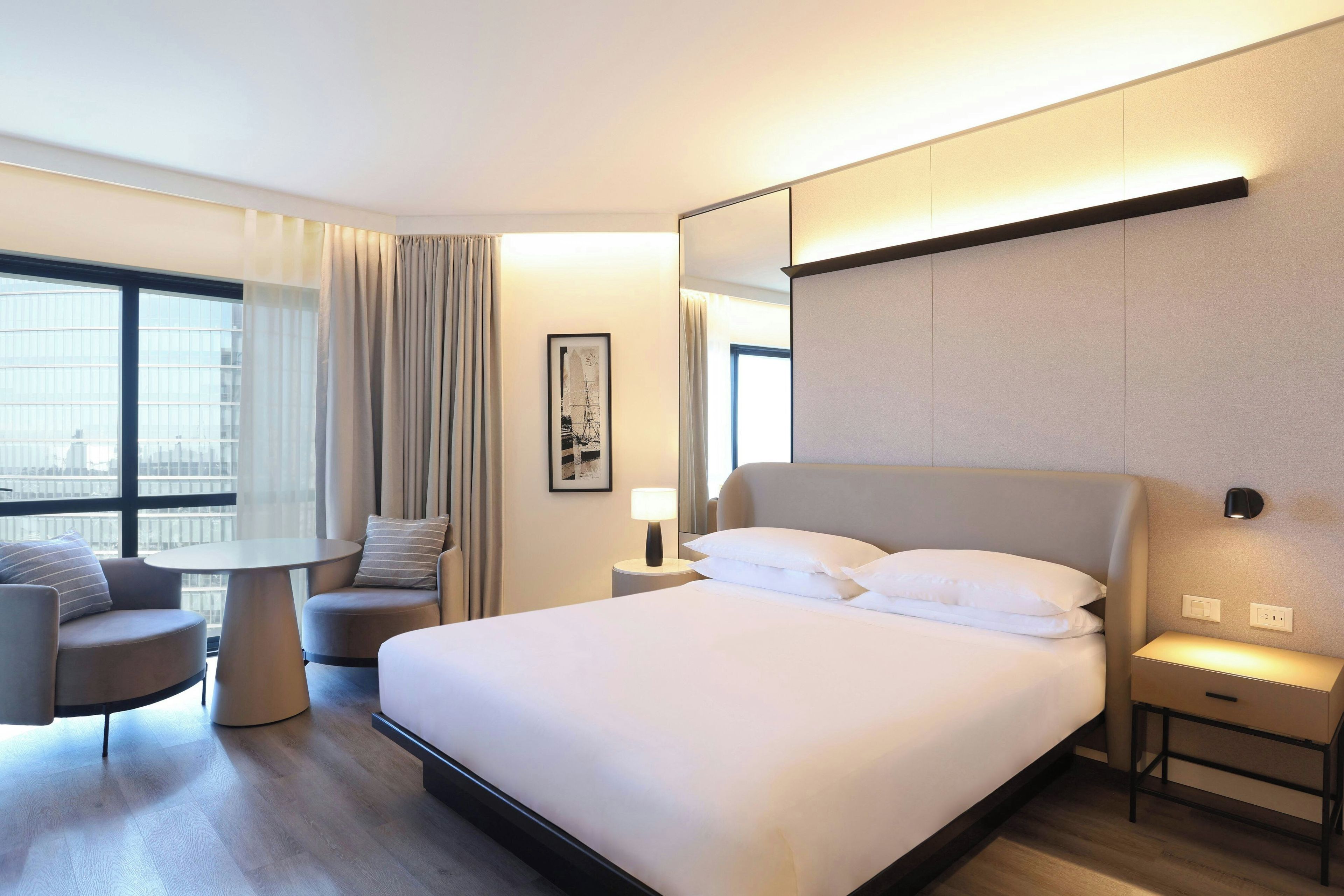 A hotel room with a large bed, a seating area with a table, and a view of the city through the window.