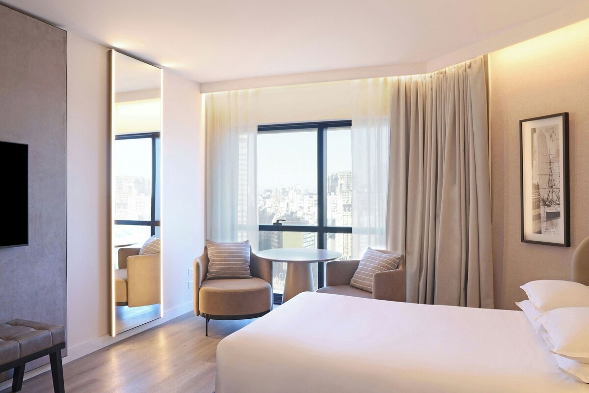 A hotel room with a bed, a round table, two armchairs, a large mirror, and a view of the city through the window.