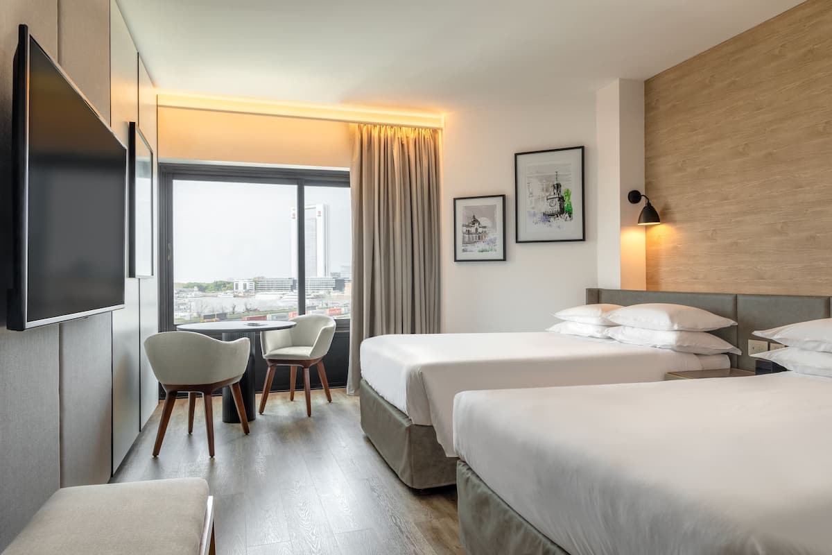 Ein Hotelzimmer mit zwei Betten, einem Schreibtisch und einem Sessel. Ein großes Fenster bietet Blick auf die Stadt.