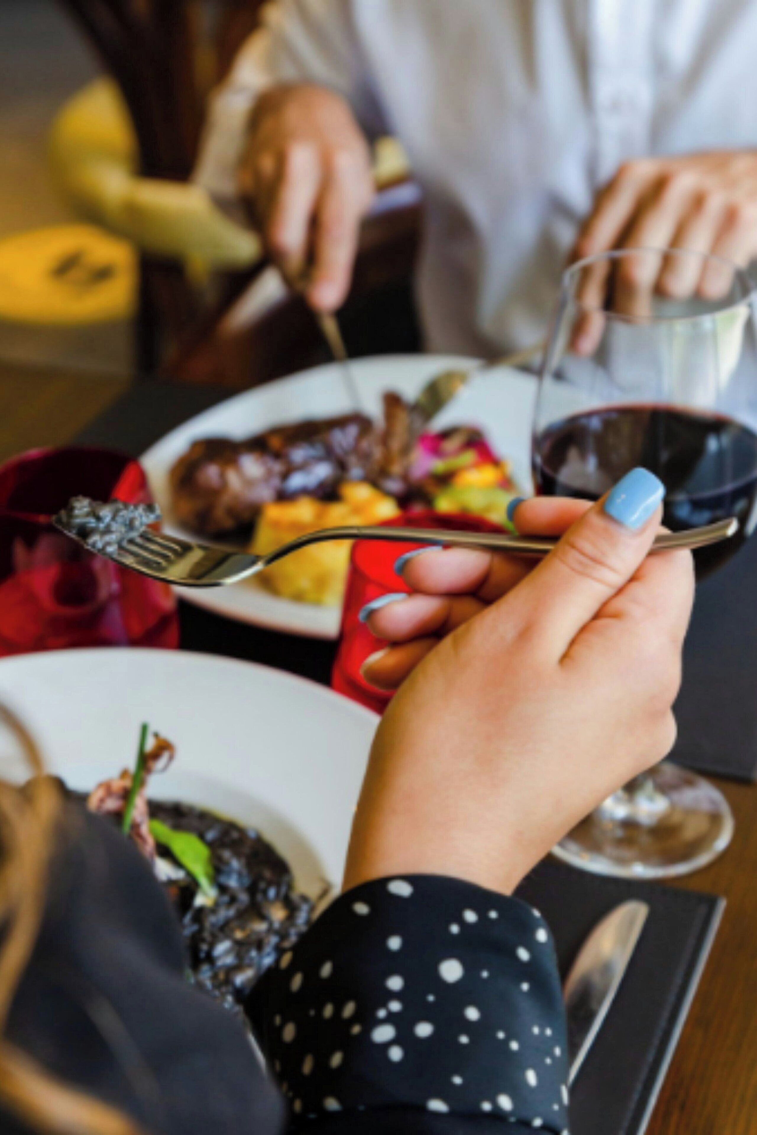 A person holding a fork and a glass of red wine, with a plate of food in the background.