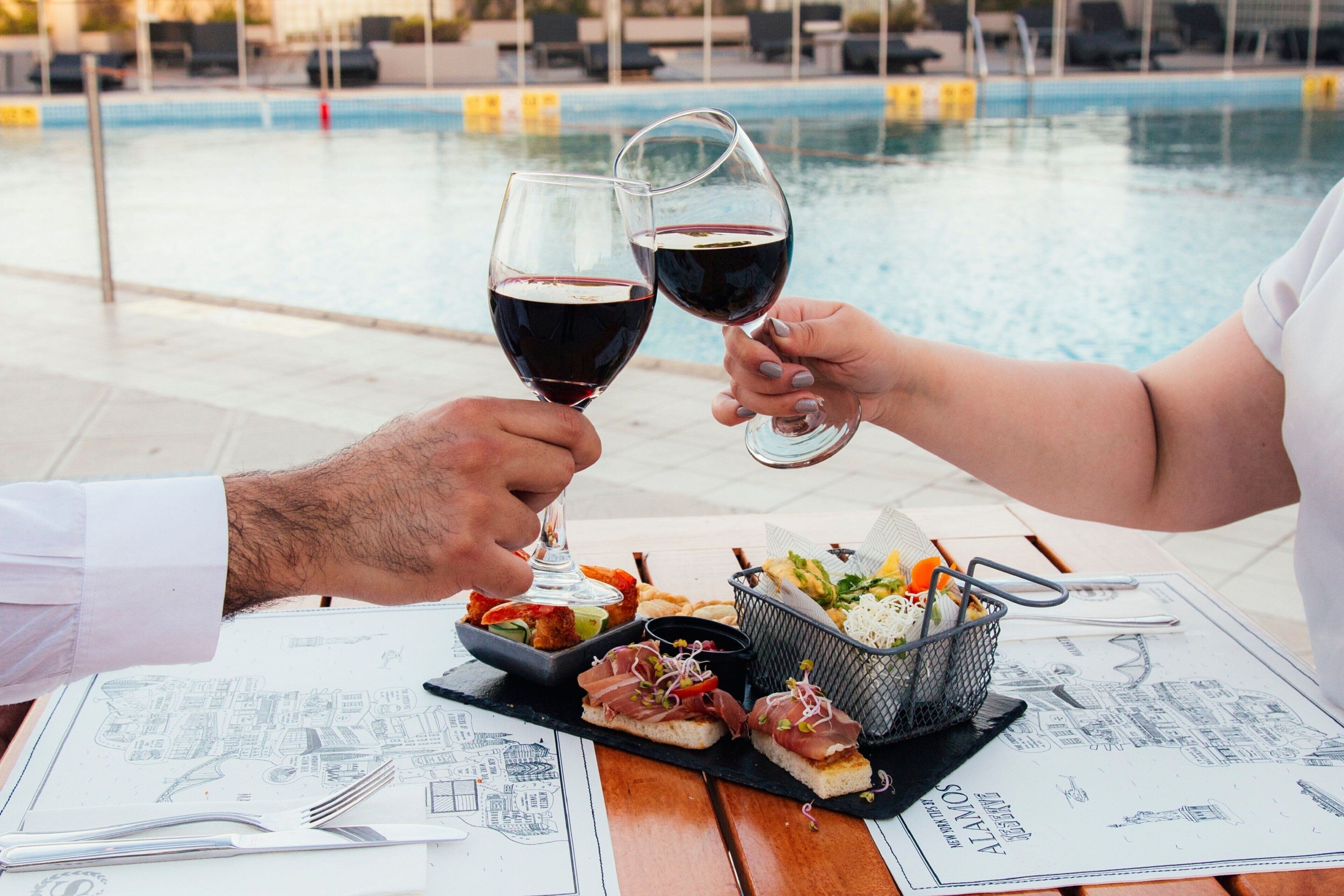 Una mesa de comedor con un menú, una piscina y dos personas brindando con copas de vino.