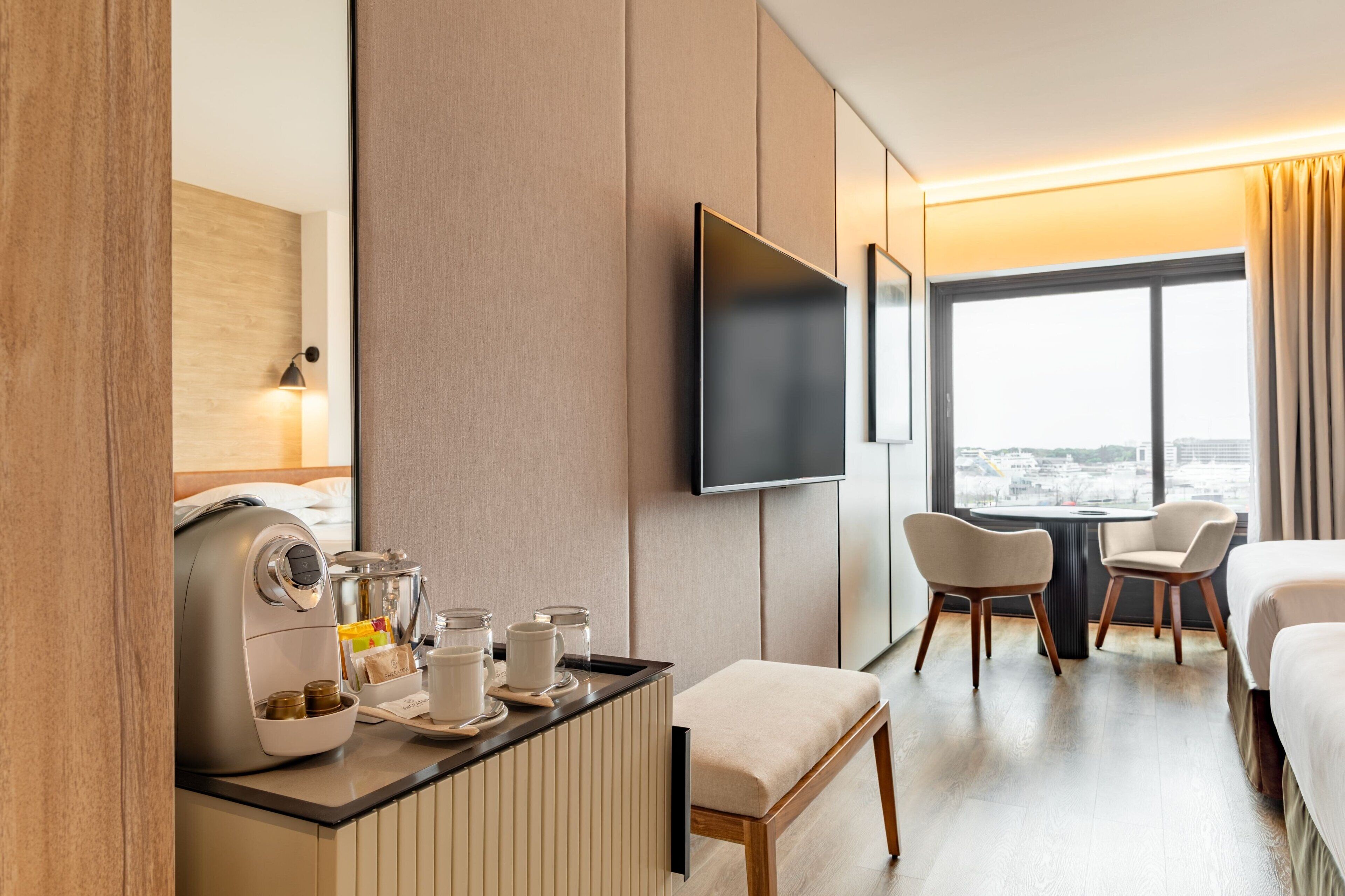 Ein Hotelzimmer mit einem Bett, einem Schreibtisch mit Kaffeemaschine, zwei Stühlen, einem Fernseher und einem großen Fenster mit Blick auf die Stadt.