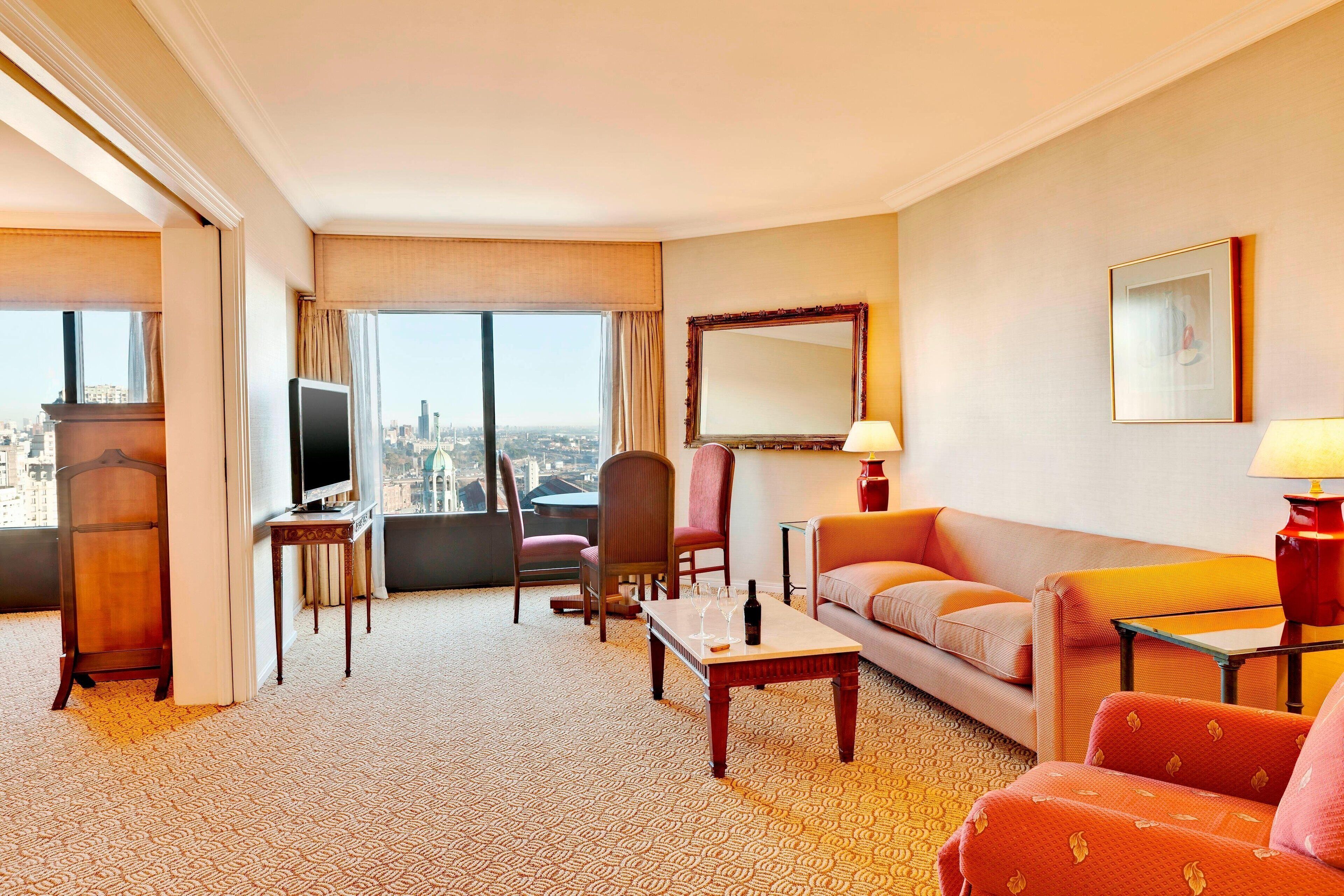 Een hotelkamer met een groot raam, een televisie, een bank, een tafel met een fles wijn en een stoel.