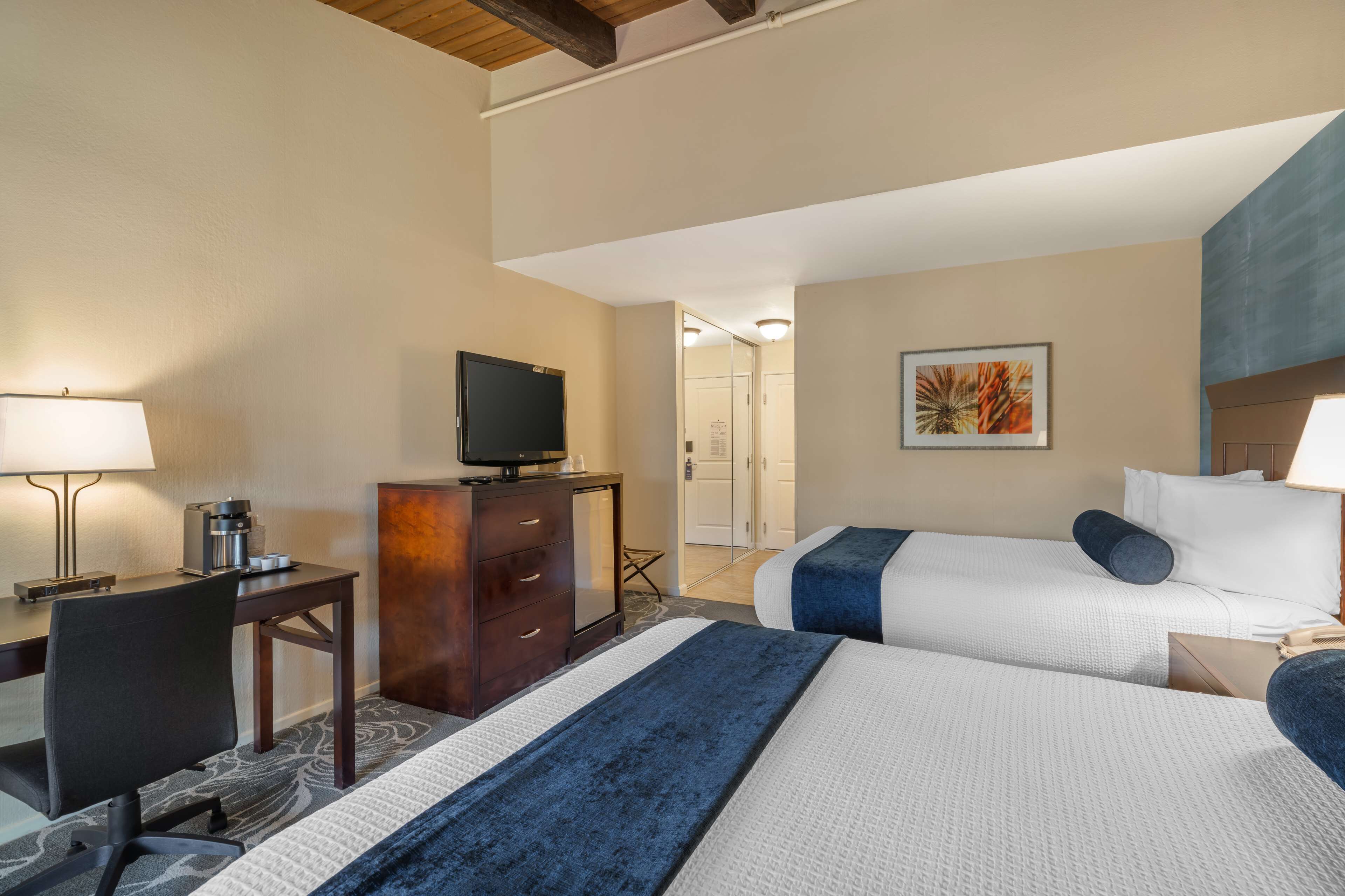 Une chambre d’hôtel avec deux lits, un bureau avec une chaise, un téléviseur posé sur une commode et un tableau accroché au mur.